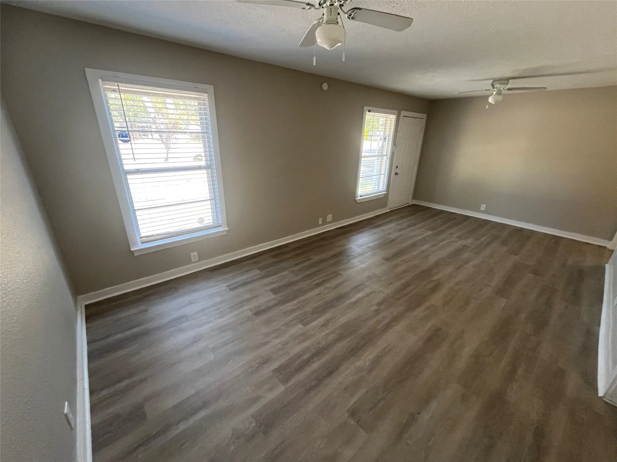 Spacious room featuring wood-finish flooring, neutral-toned walls, multiple windows with blinds, and two ceiling fans