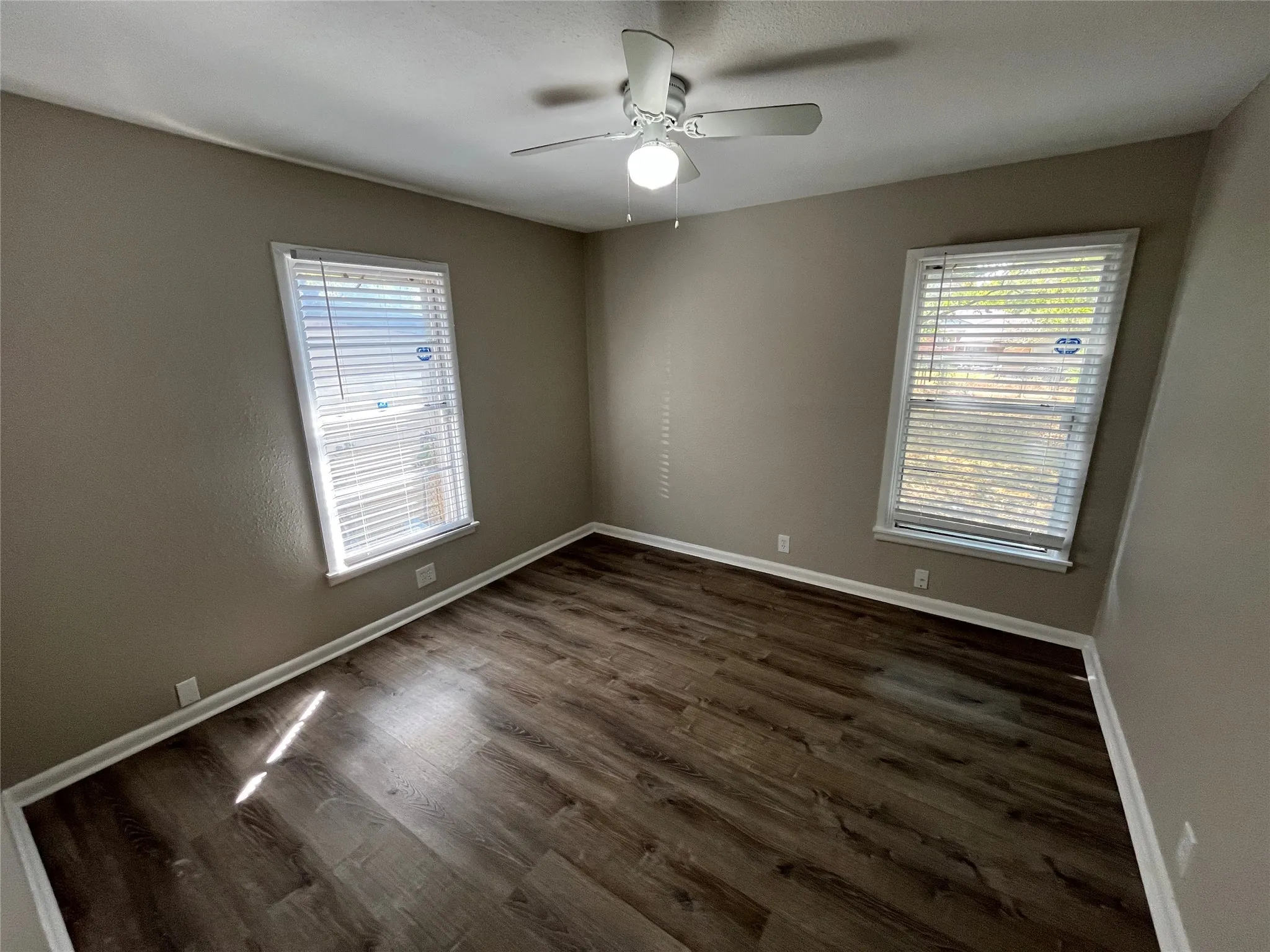 Wood-finish flooring, neutral wall tones, white trim, two windows with blinds, and a ceiling fan with integrated lighting