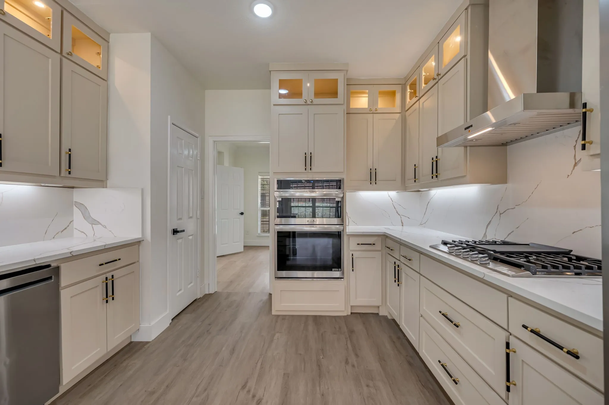 Gourmet kitchen featuring a built-in double oven, gas cooktop, stainless steel range hood, stone countertops with matching backsplash, and custom cabinetry with glass-front uppers