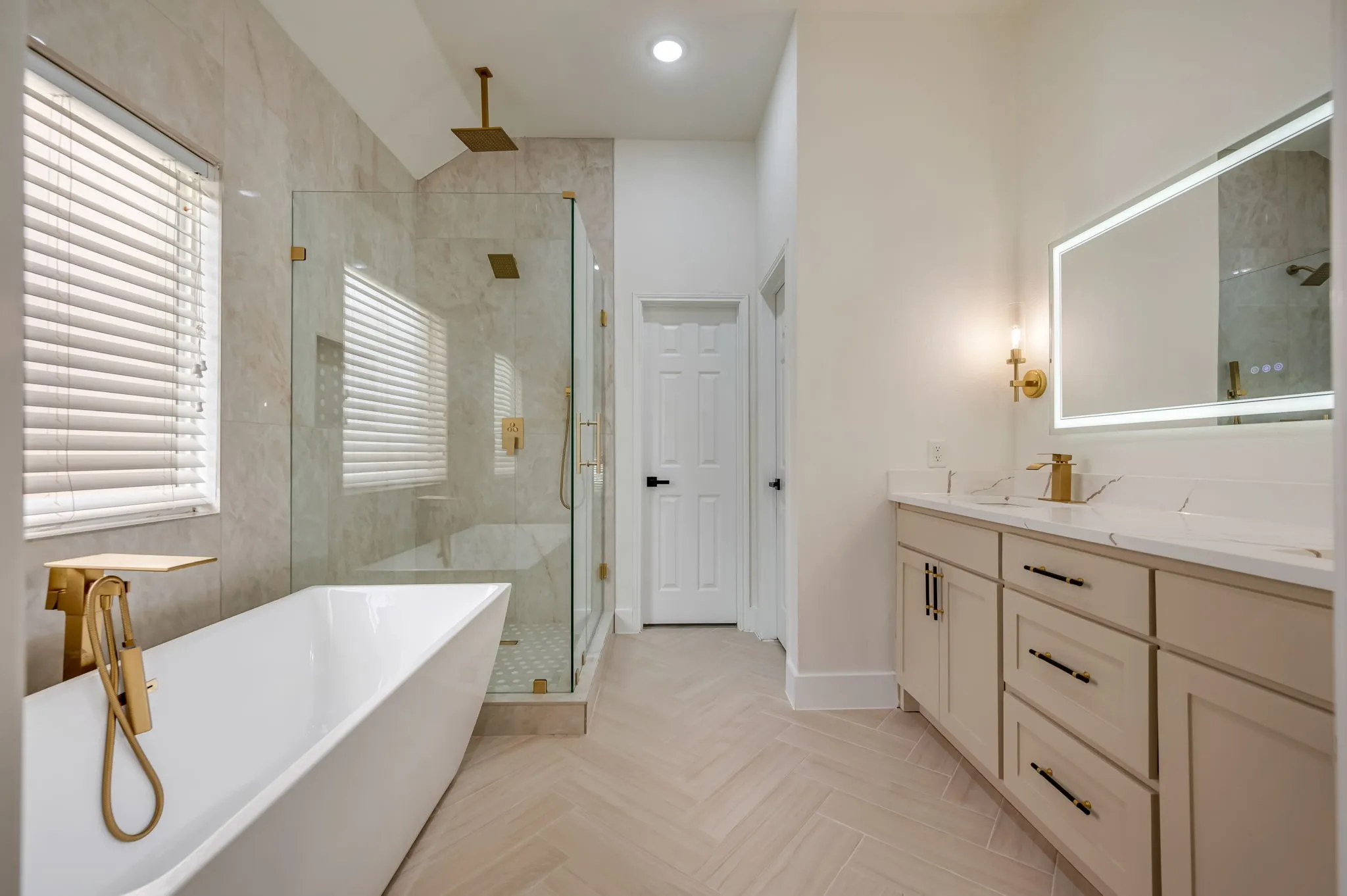 Freestanding soaking tub with gold-tone fixtures, walk-in shower with glass enclosure, dual vanity with undermount sinks, illuminated mirror, and herringbone-patterned tile flooring