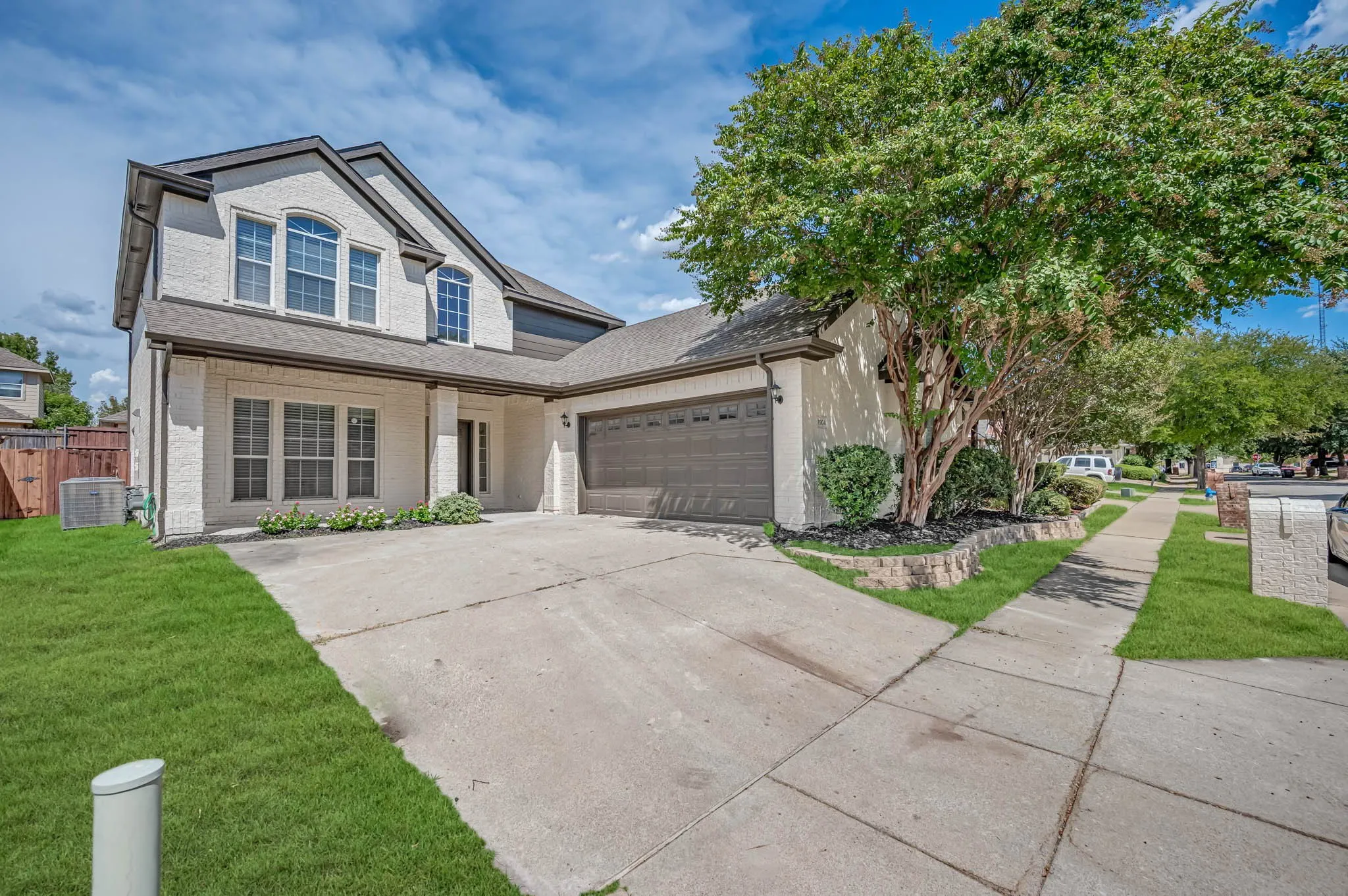 Two-story exterior with Beige brick, dark trim, a two-car garage, established landscaping, and a concrete driveway