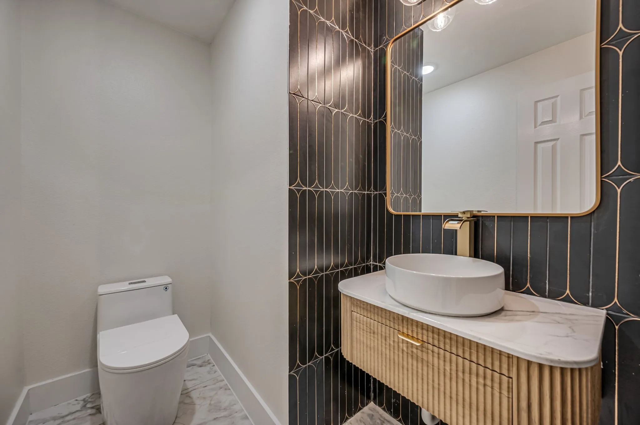 Powder room featuring a sleek toilet, patterned wall tile, gold-framed mirror, floating vanity with ribbed Luxury Vinyl Plank  cabinetry, and a countertop vessel sink with a gold-tone faucet