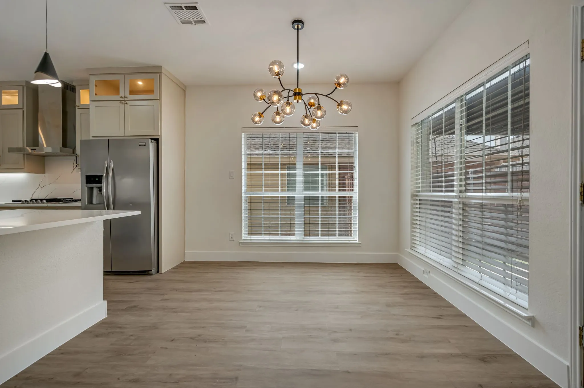 Open-concept living space featuring Luxury Vinyl Plank  flooring, a contemporary chandelier, large windows with blinds, and a kitchen with stainless steel appliances and light-toned cabinetry