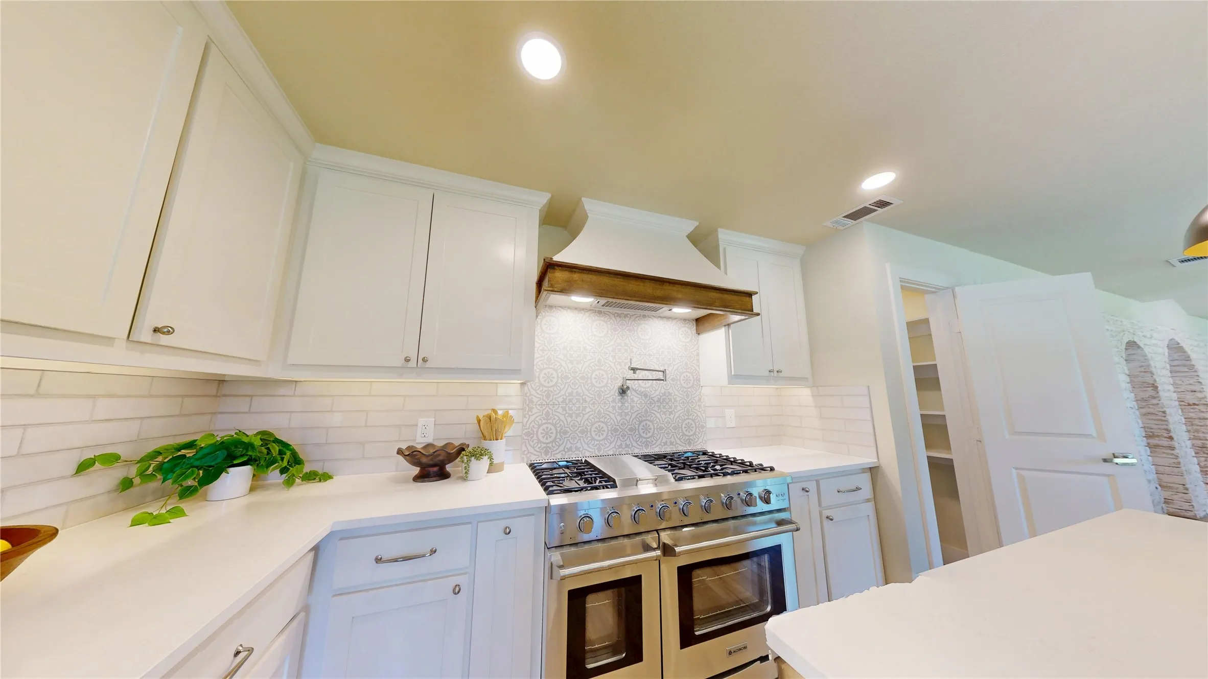 Stainless steel range with double ovens, custom wood-trimmed range hood, mosaic tile backsplash, white shaker cabinetry, and a walk-in pantry