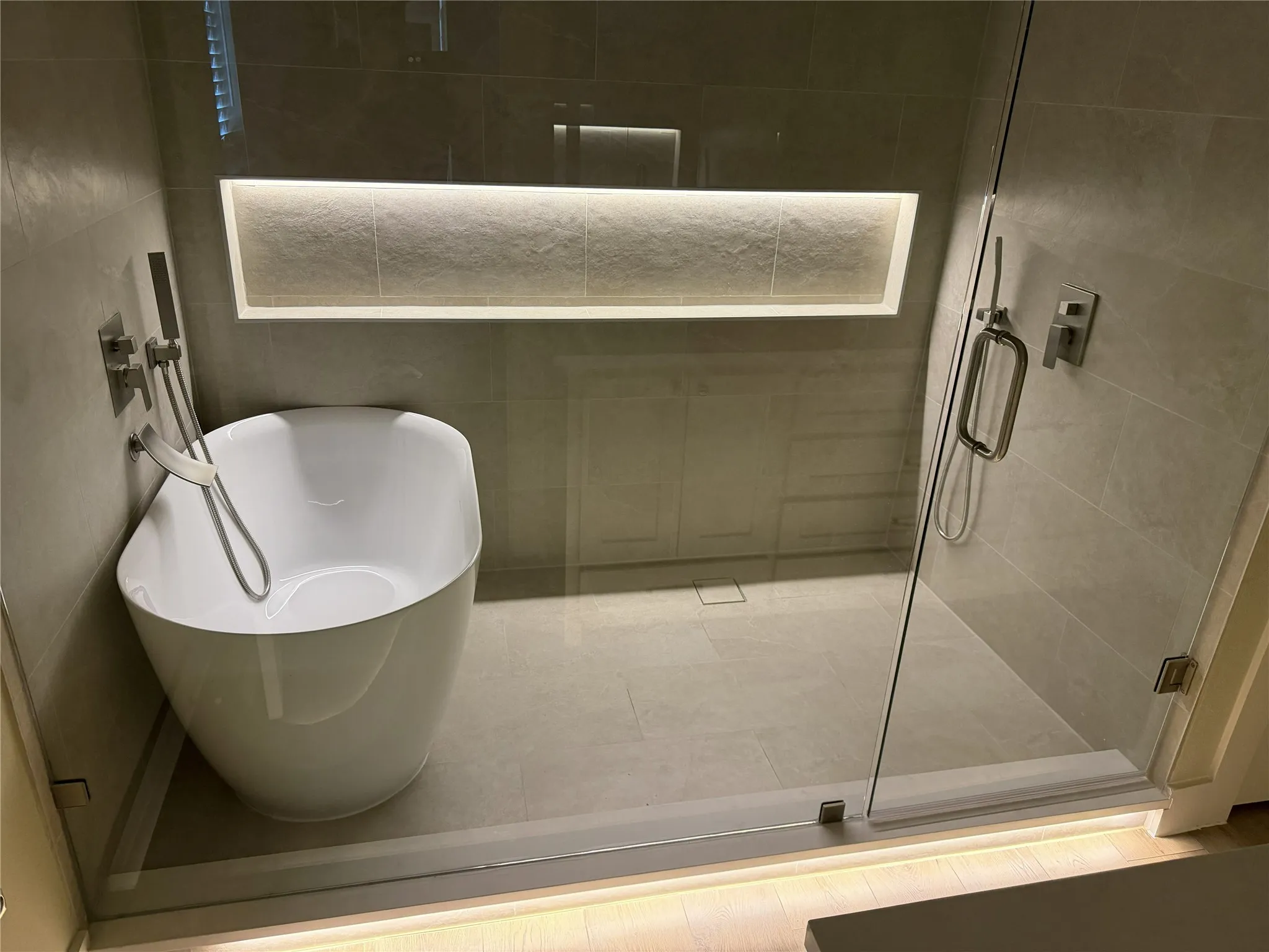 Freestanding soaking tub with a wall-mounted faucet, integrated shower niche with accent lighting, glass-enclosed shower, and tiled flooring
