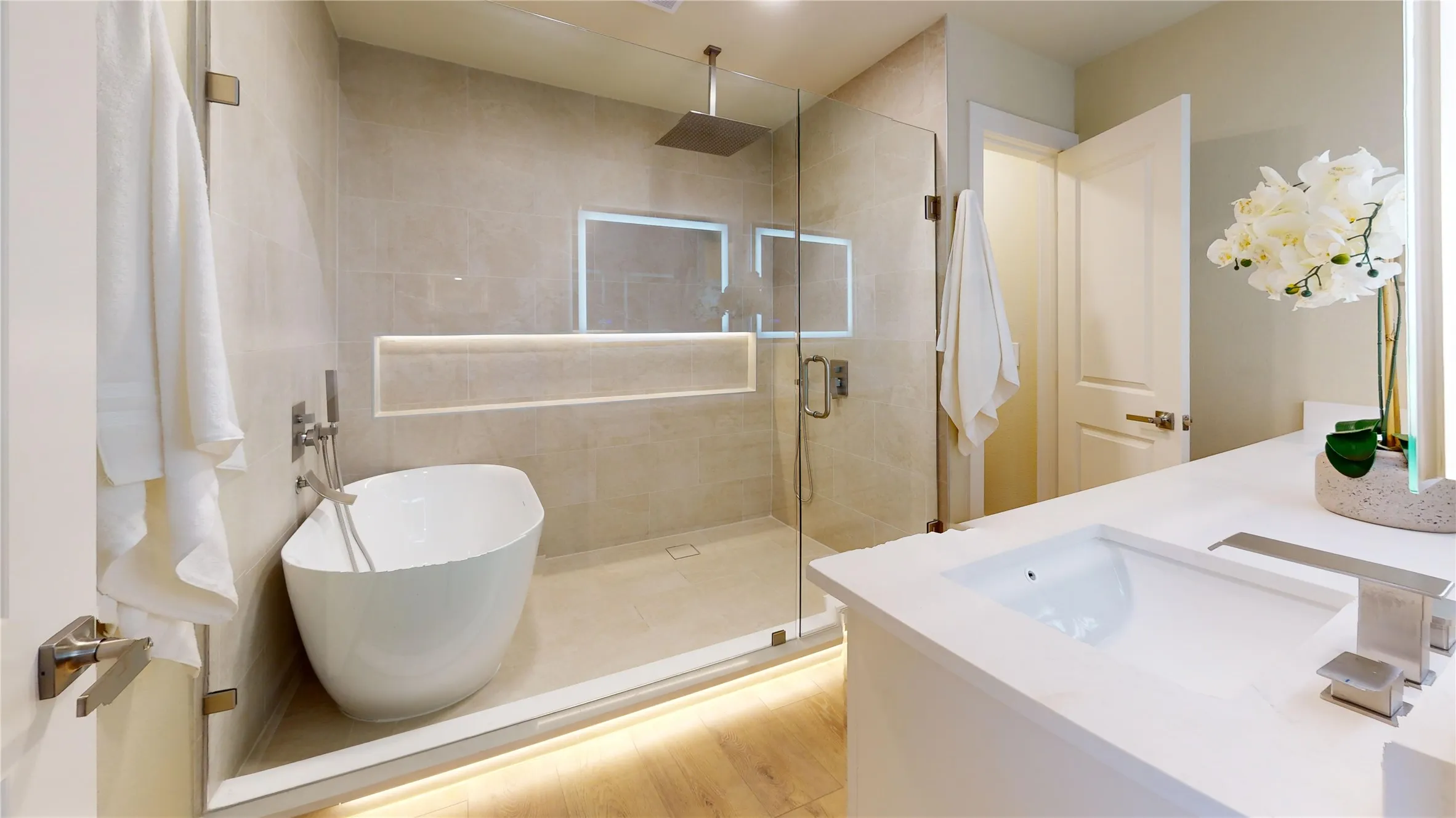 Freestanding soaking tub, glass-enclosed shower with rainhead, integrated shower niche, floating vanity with integrated sink, and wood-finish flooring