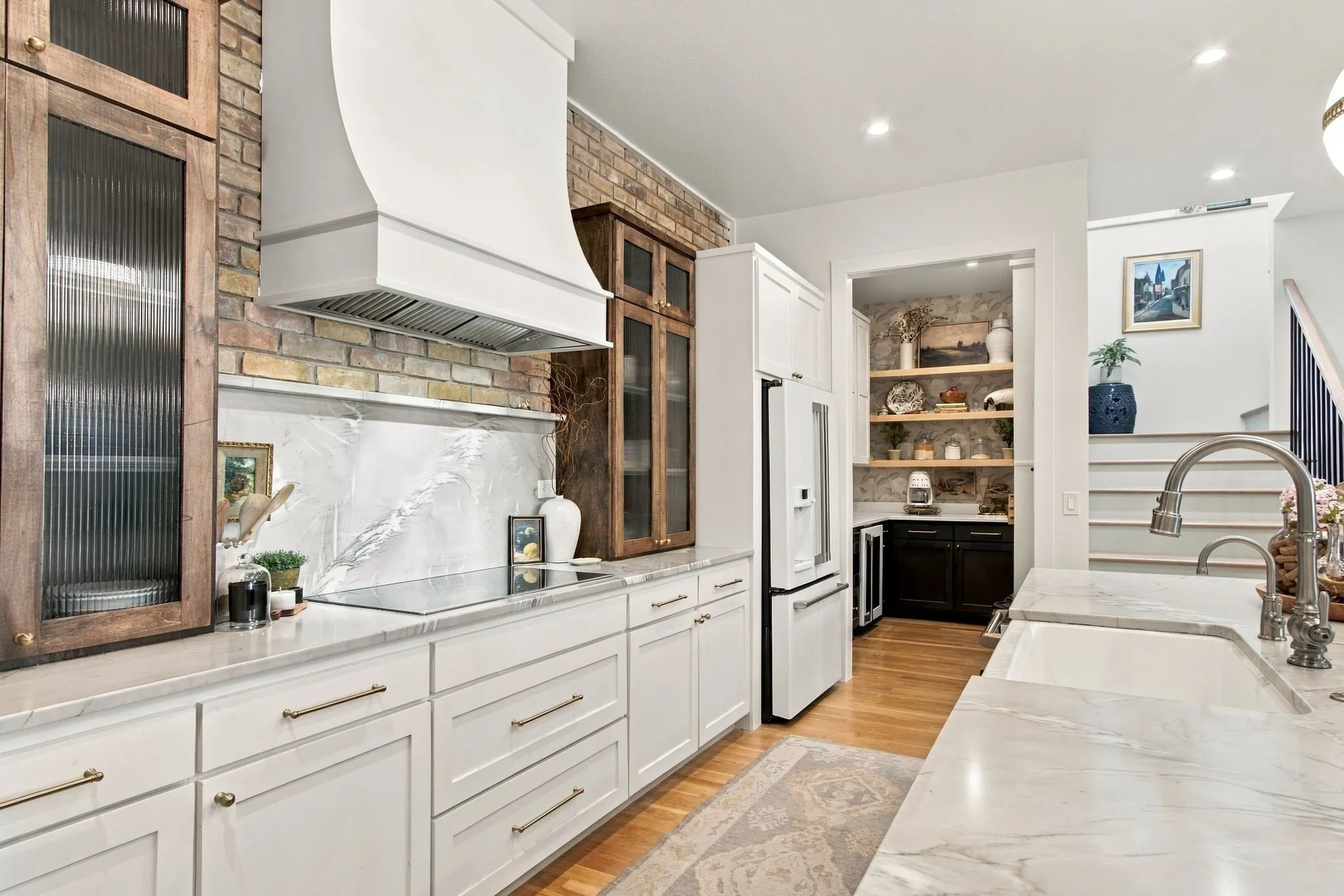 White shaker cabinetry with brushed brass hardware, brick backsplash, integrated range hood, under-counter beverage refrigerator, and a built-in kitchen sink