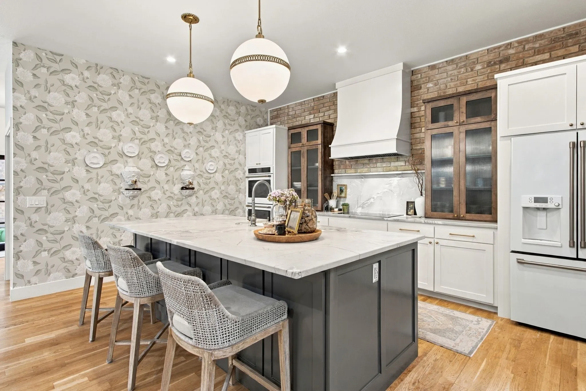 Spacious kitchen island with a light-toned countertop, wood-finish flooring, two spherical pendant lights, exposed brick accent wall, and integrated stainless steel appliances