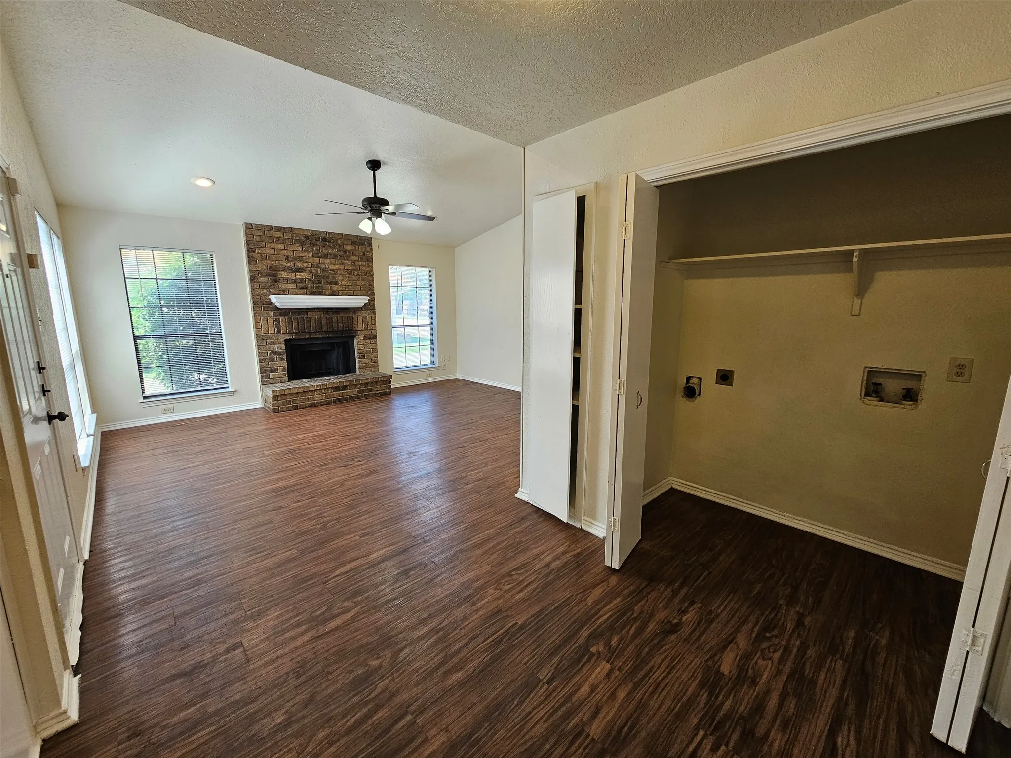 Vaulted ceiling, wood-finish flooring, brick fireplace with a white mantel, ceiling fan, and built-in laundry closet with shelving