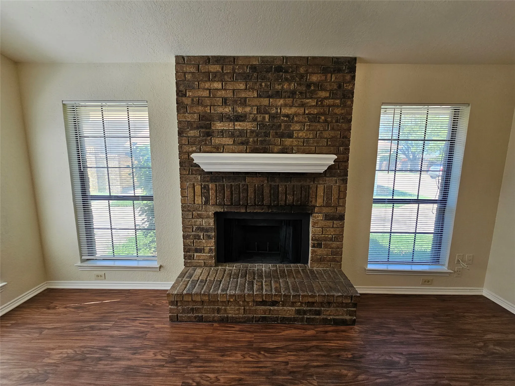 Brick fireplace with a classic white mantel, flanked by two windows with horizontal blinds, and wood-finish flooring