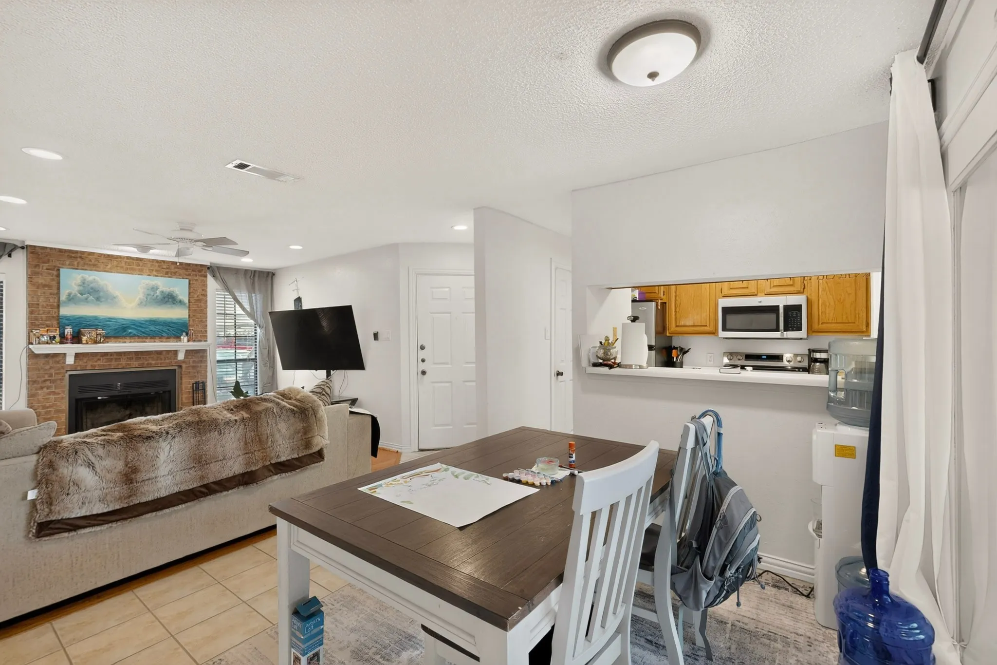 Brick-faced fireplace with a mantel, tile flooring, white walls, wood-finish dining table, and a kitchen pass-through with wood cabinetry