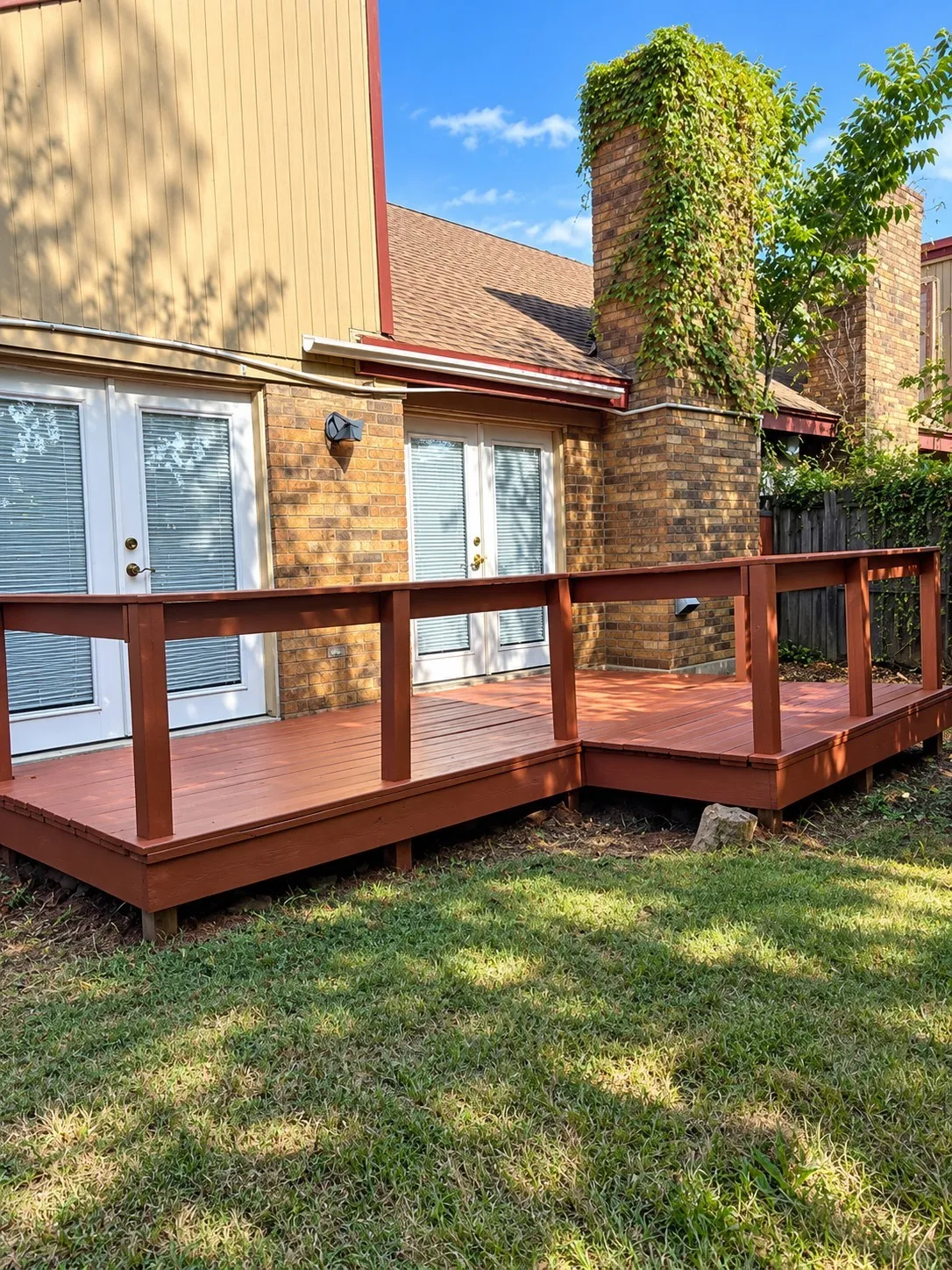 Multi-level wood deck with protective railing, two sets of French doors, brick chimney with natural climbing foliage, and a landscaped lawn area