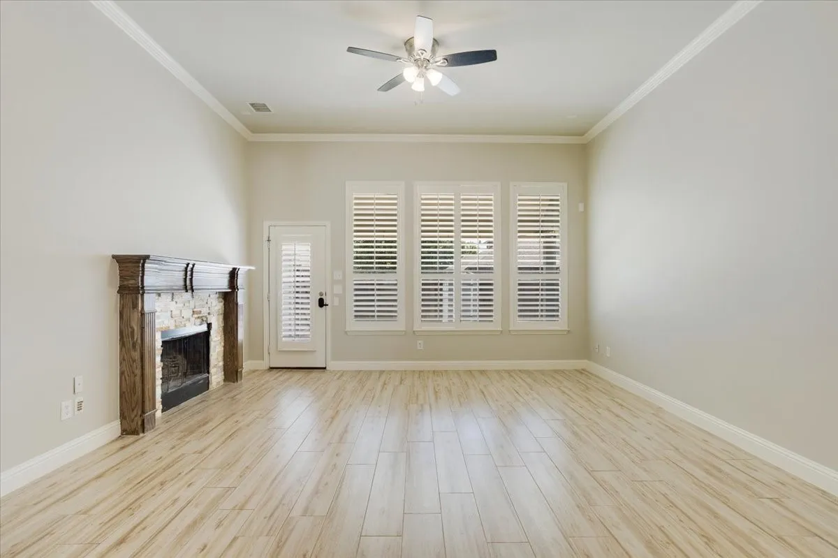 Spacious living area featuring wood-finish flooring, a stone-accented fireplace with a wood mantle, custom plantation shutters, and crown molding