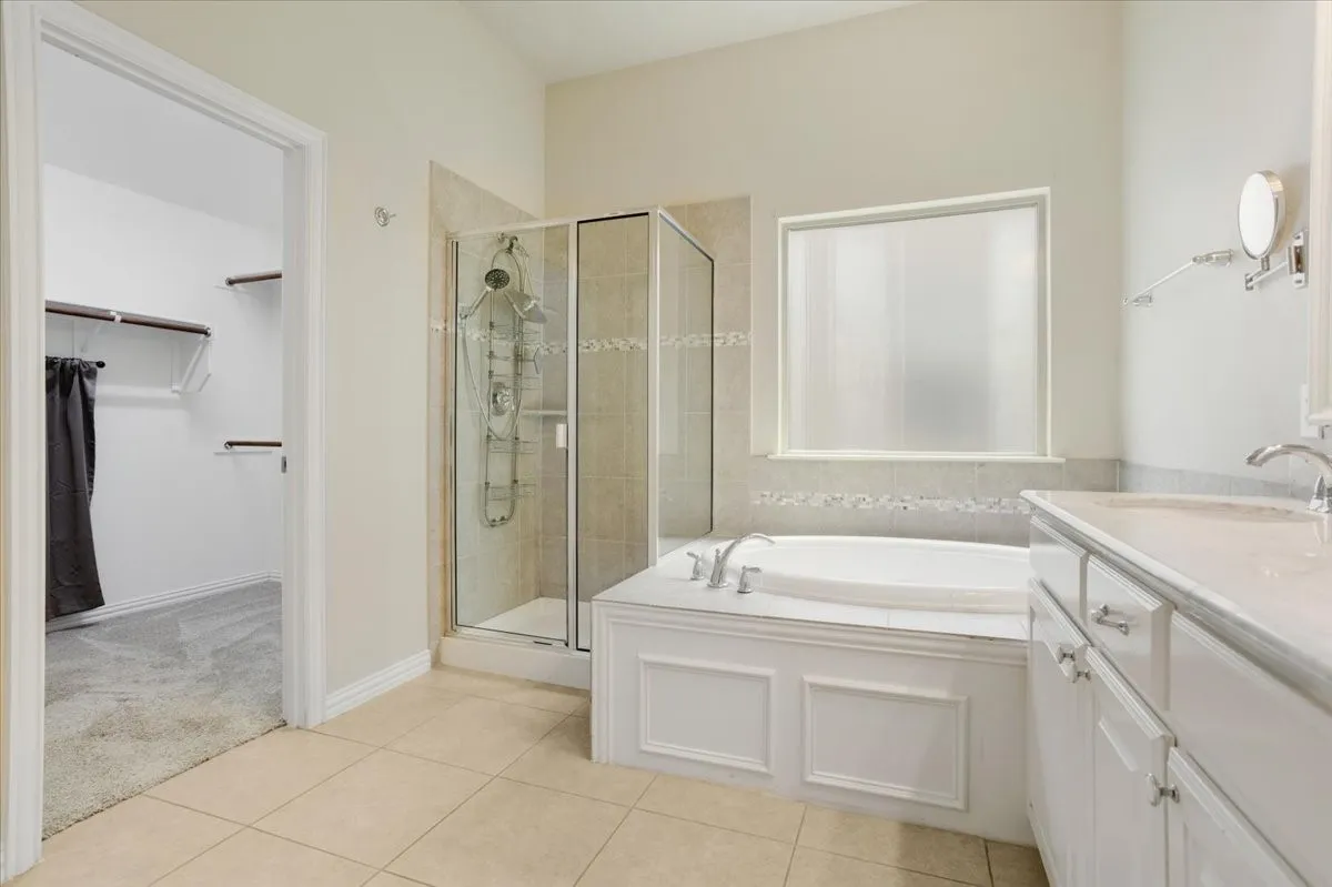 Tiled flooring throughout, a built-in tub with paneling, a glass-enclosed shower, dual vanity with a light-colored countertop, and a walk-in closet with built-in shelving