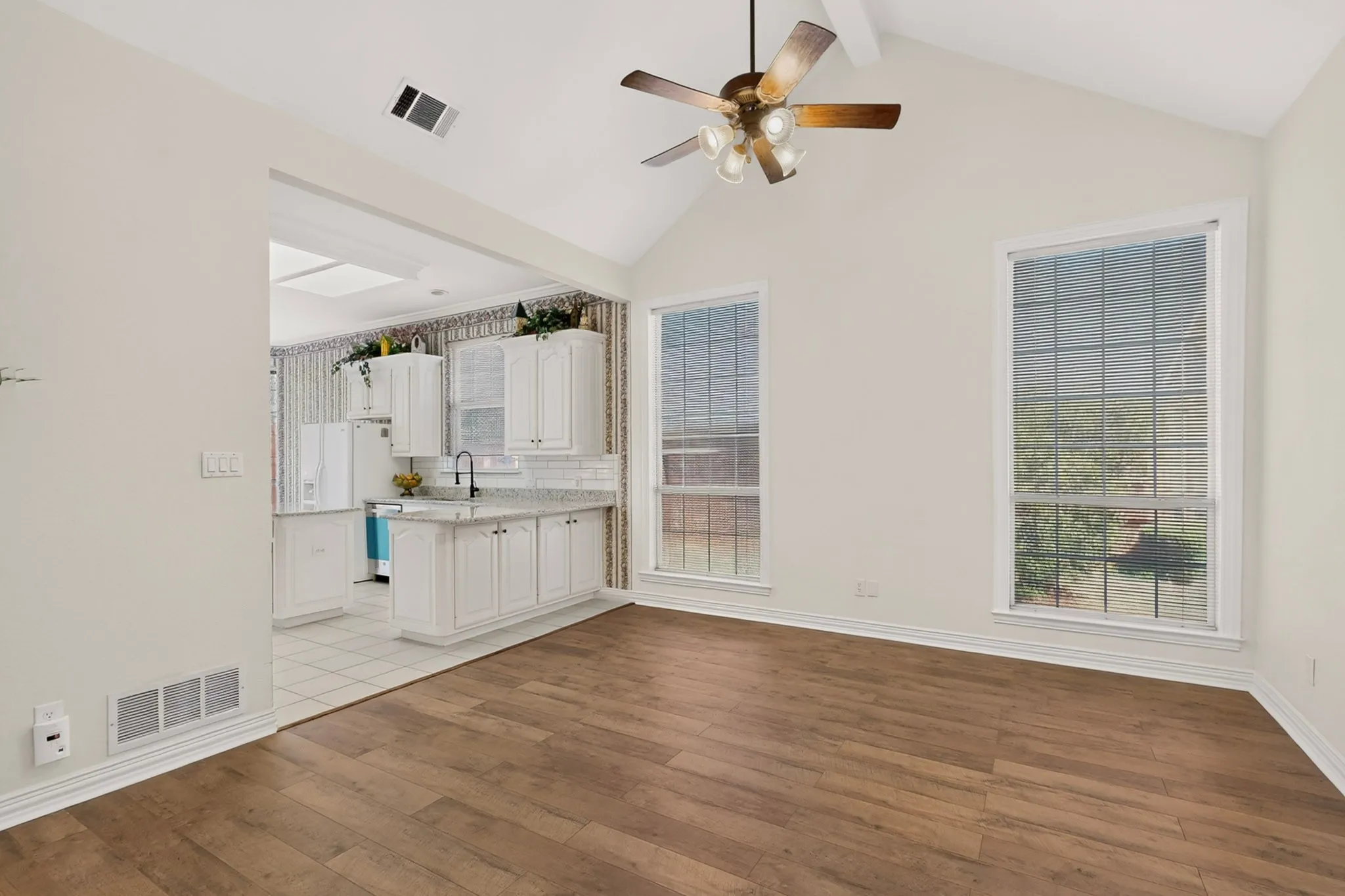 Vaulted ceiling with a ceiling fan, wood-finish flooring, two large windows, white cabinetry, and a subway tile backsplash