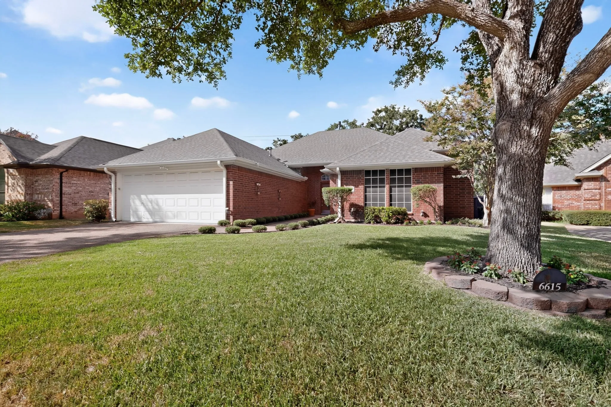 Brick exterior featuring a two-car garage, pitched shingle roof, and established landscaping