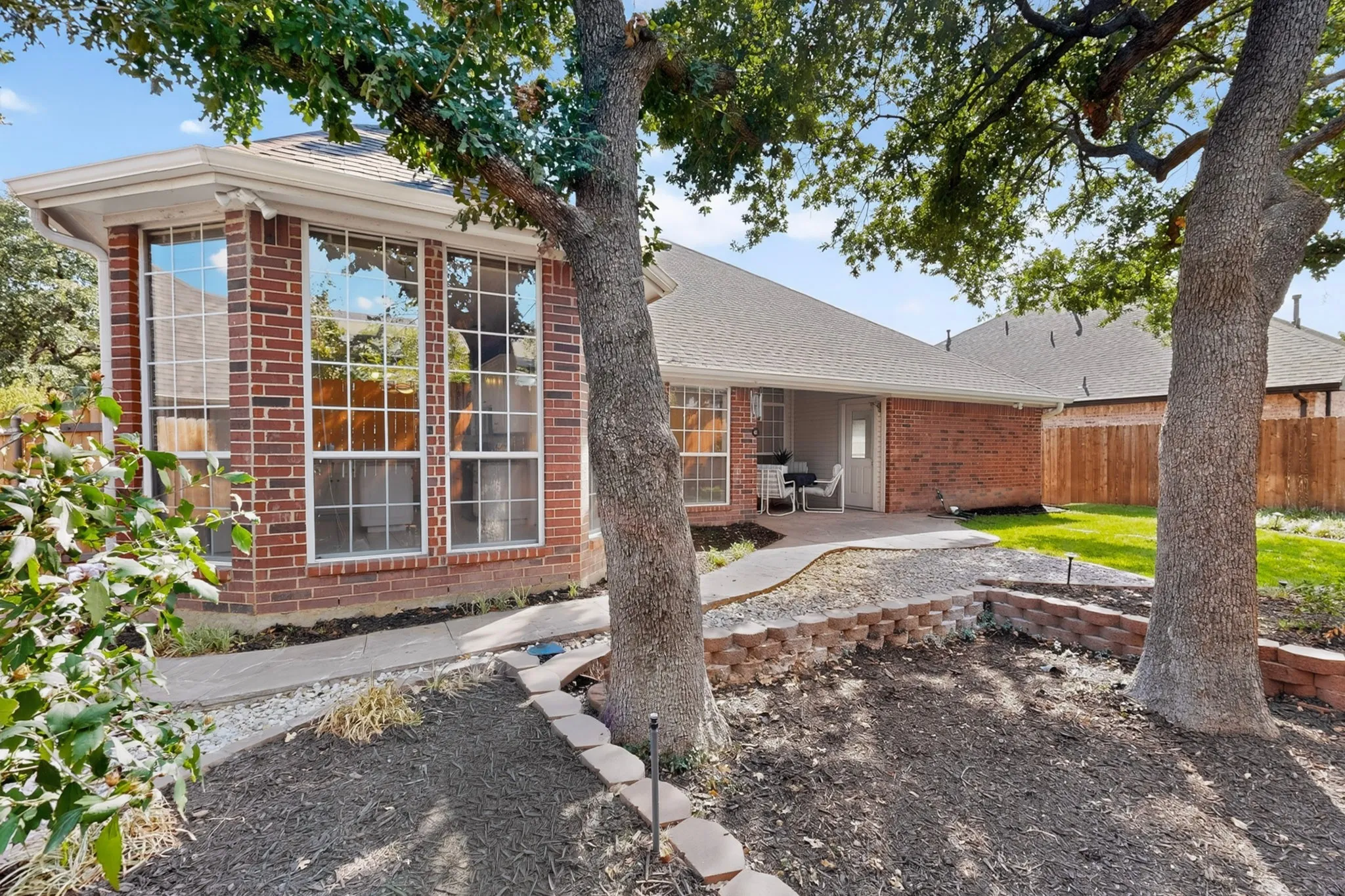 Brick exterior with a covered patio, expansive windows, mature trees, and a well-maintained lawn