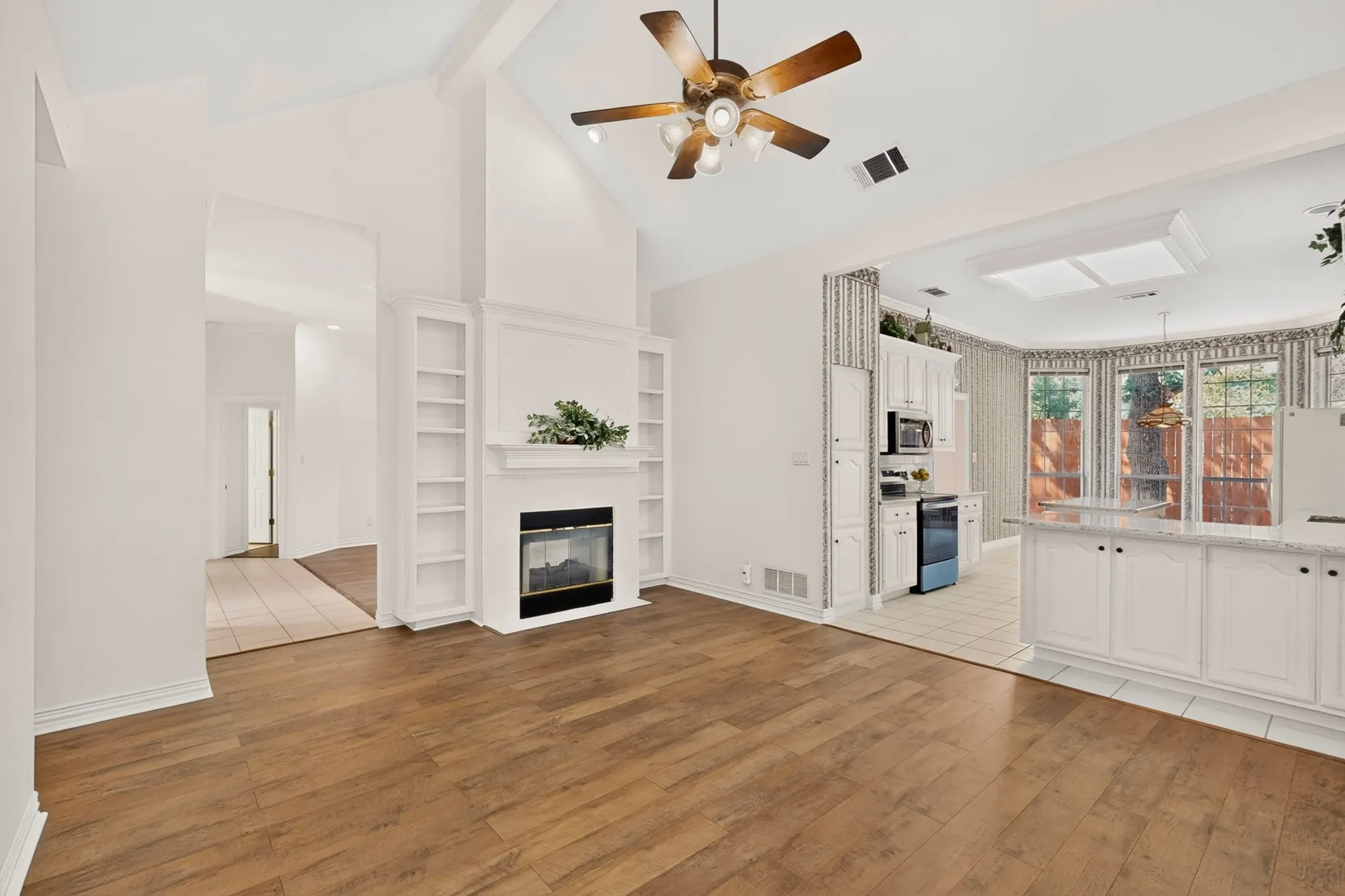 Vaulted ceiling with a ceiling fan, fireplace with integrated built-in shelving, wood-finish flooring, and a kitchen with white cabinetry and a skylight