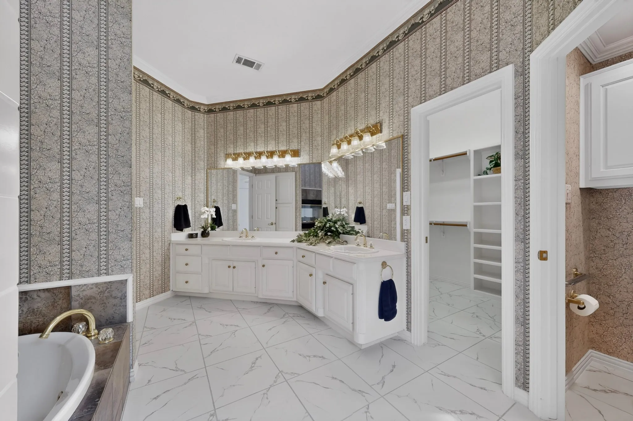 Double vanity with integrated sinks and white cabinetry, large vanity mirror, built-in bathtub with brass fixtures, and a walk-in closet with shelving and hanging rods