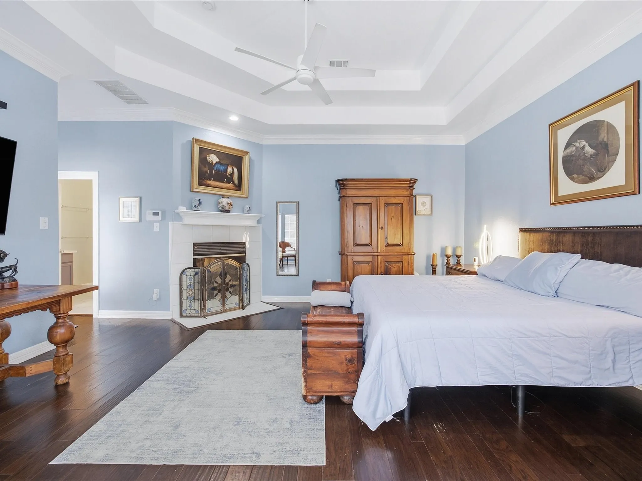 Tray ceiling with a ceiling fan, wood-finish flooring, white mantel fireplace, and light blue walls