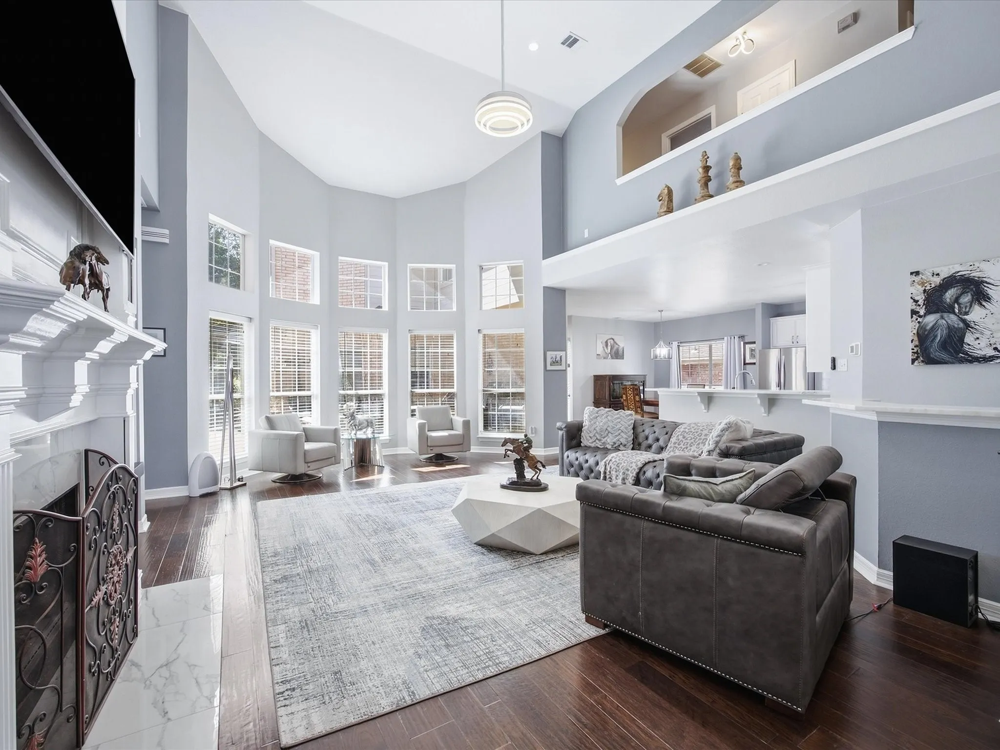 Vaulted ceiling with a contemporary light fixture, wood-finish flooring, a white mantel fireplace, multiple large windows, and an upper display ledge
