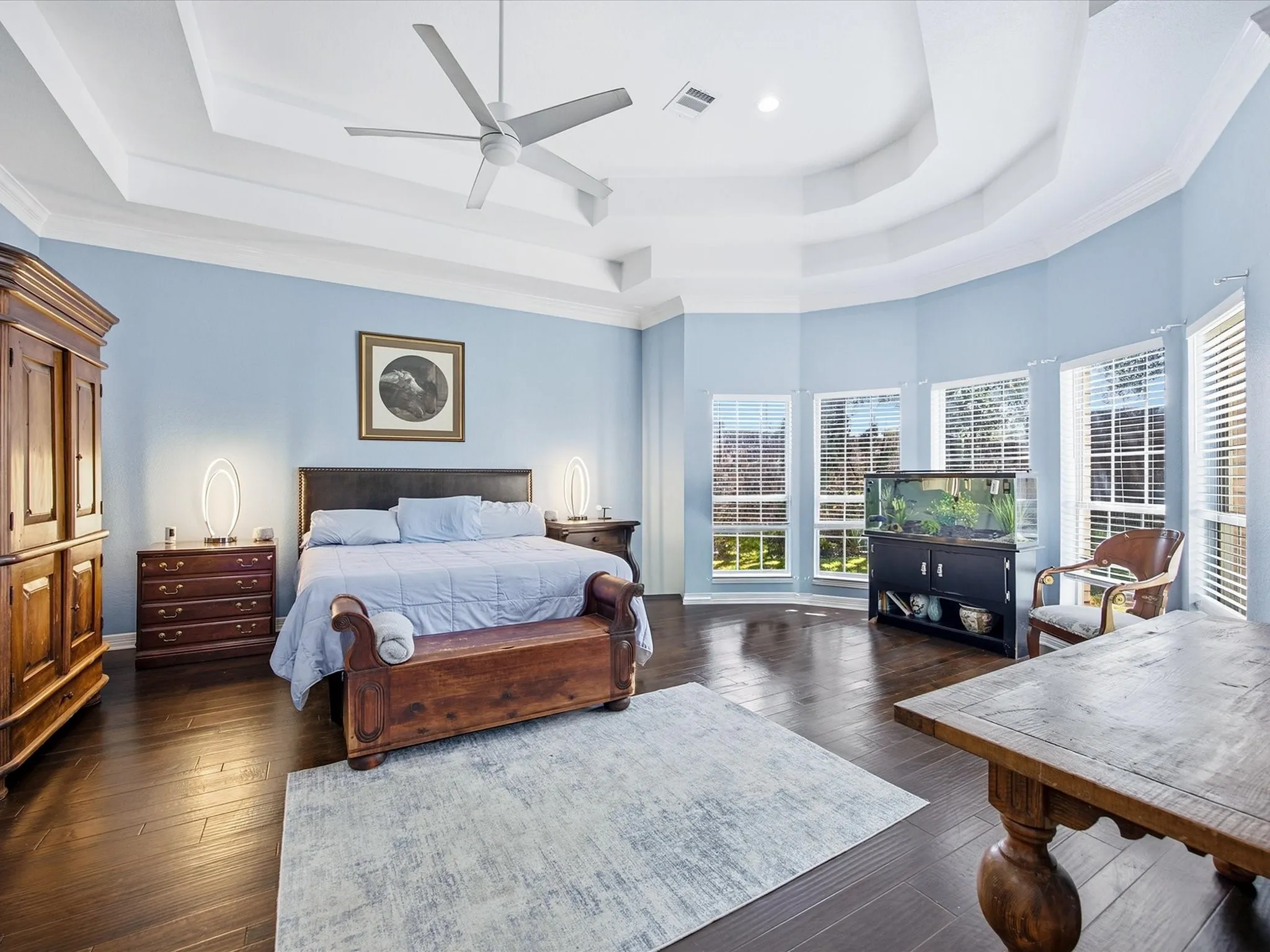 Tray ceiling with crown molding, ceiling fan, wood-finish flooring, and multiple windows with blinds