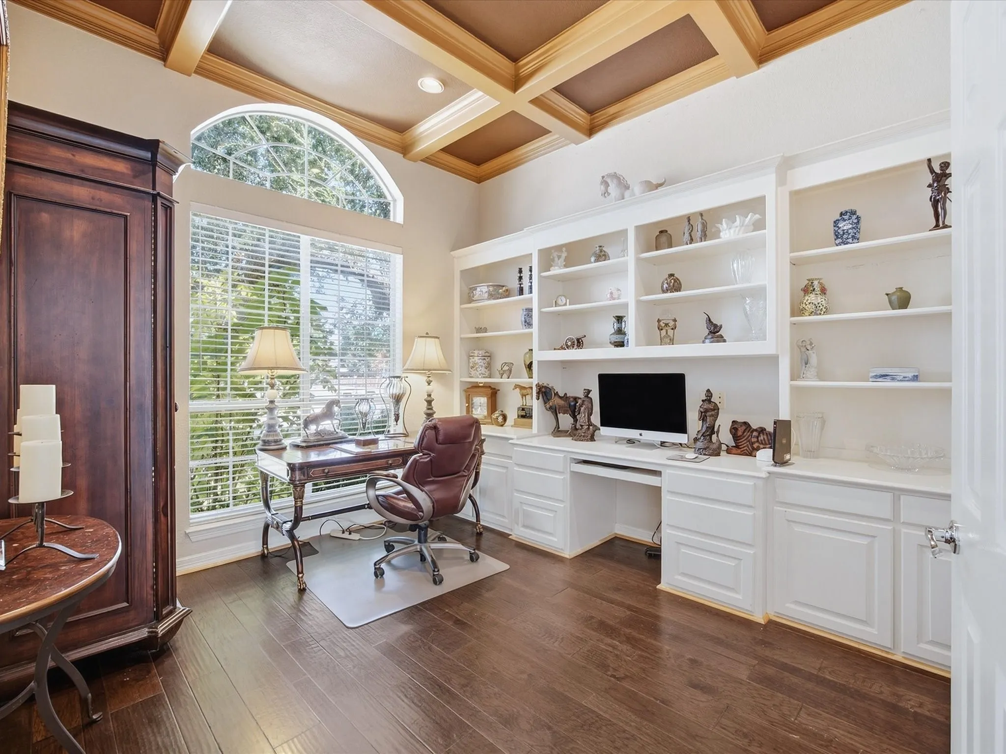 Built-in white shelving and cabinetry, coffered ceiling, dark wood-finish flooring, and a large arched window