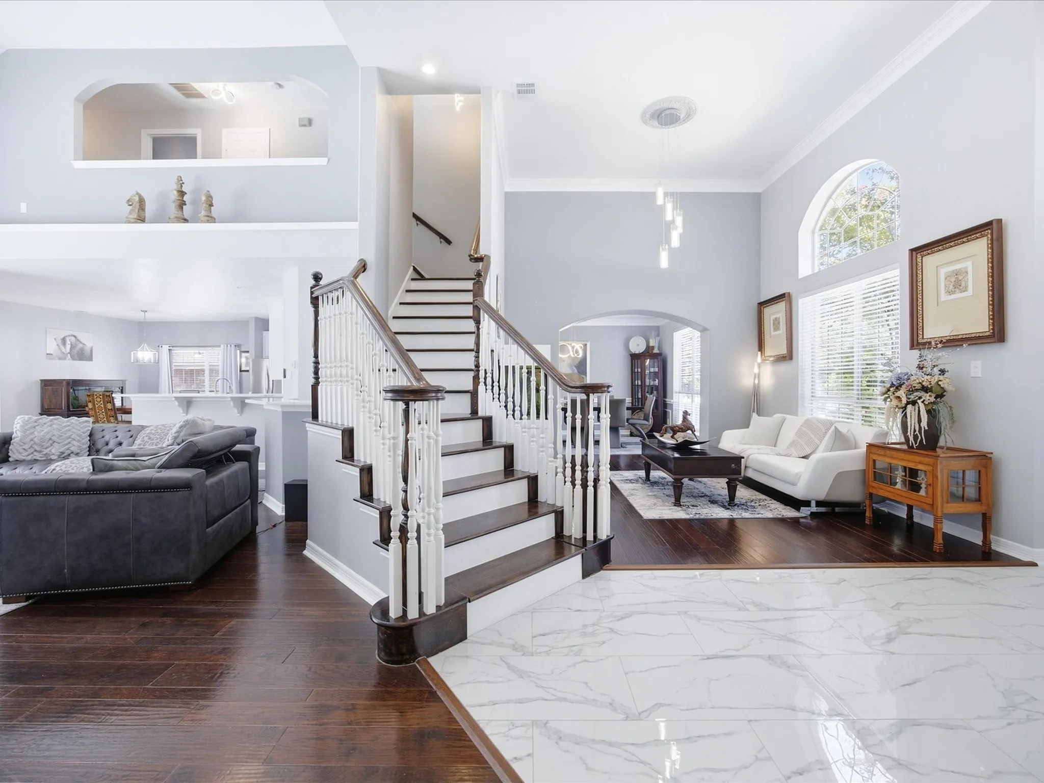 Grand foyer featuring high ceilings, a prominent staircase with dark wood-finish treads and white balusters, large arched windows, a modern entry door, and polished marble-look flooring