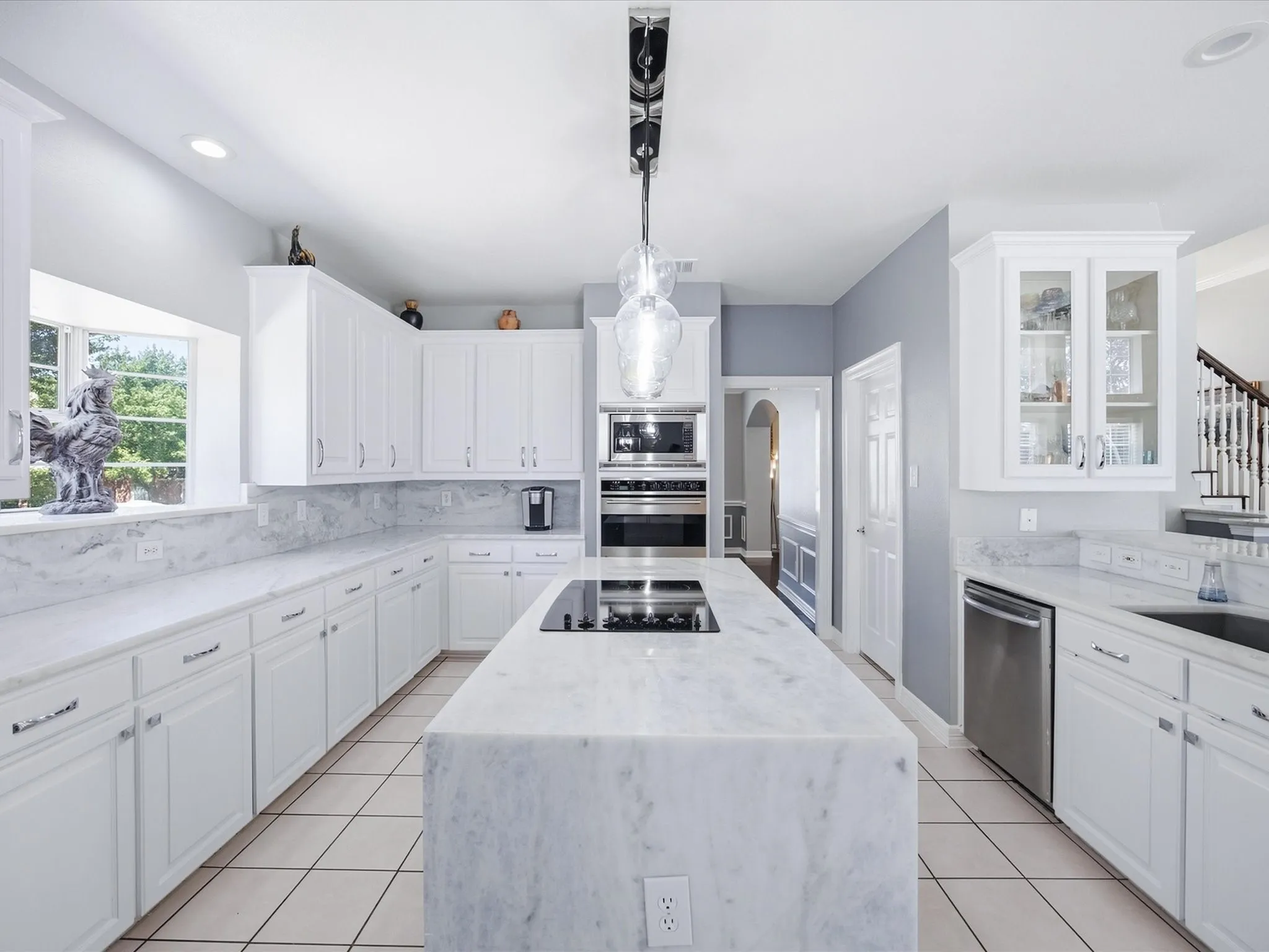 White cabinetry with glass-front accents, integrated wall oven and microwave, stainless steel dishwasher, large center island with cooktop, and tile flooring