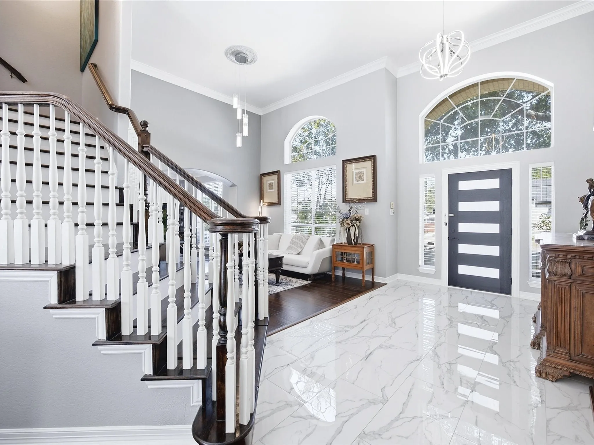 Grand foyer featuring high ceilings, a prominent staircase with dark wood-finish treads and white balusters, large arched windows, a modern entry door, and polished marble-look flooring