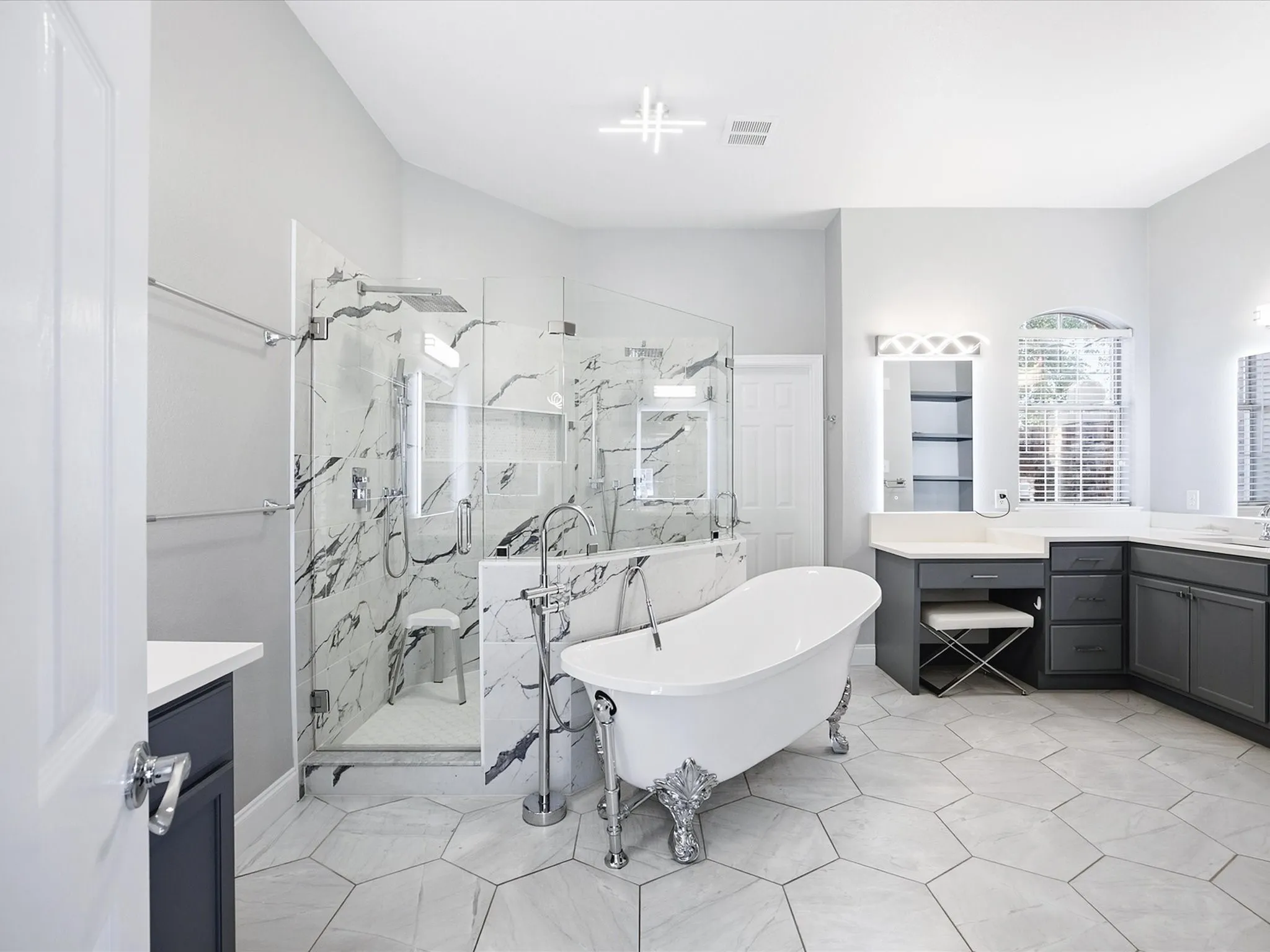 Freestanding clawfoot tub, glass-enclosed shower with marble-look tile surround, hexagonal floor tile, dual vanity with undermount sinks, and stone countertops