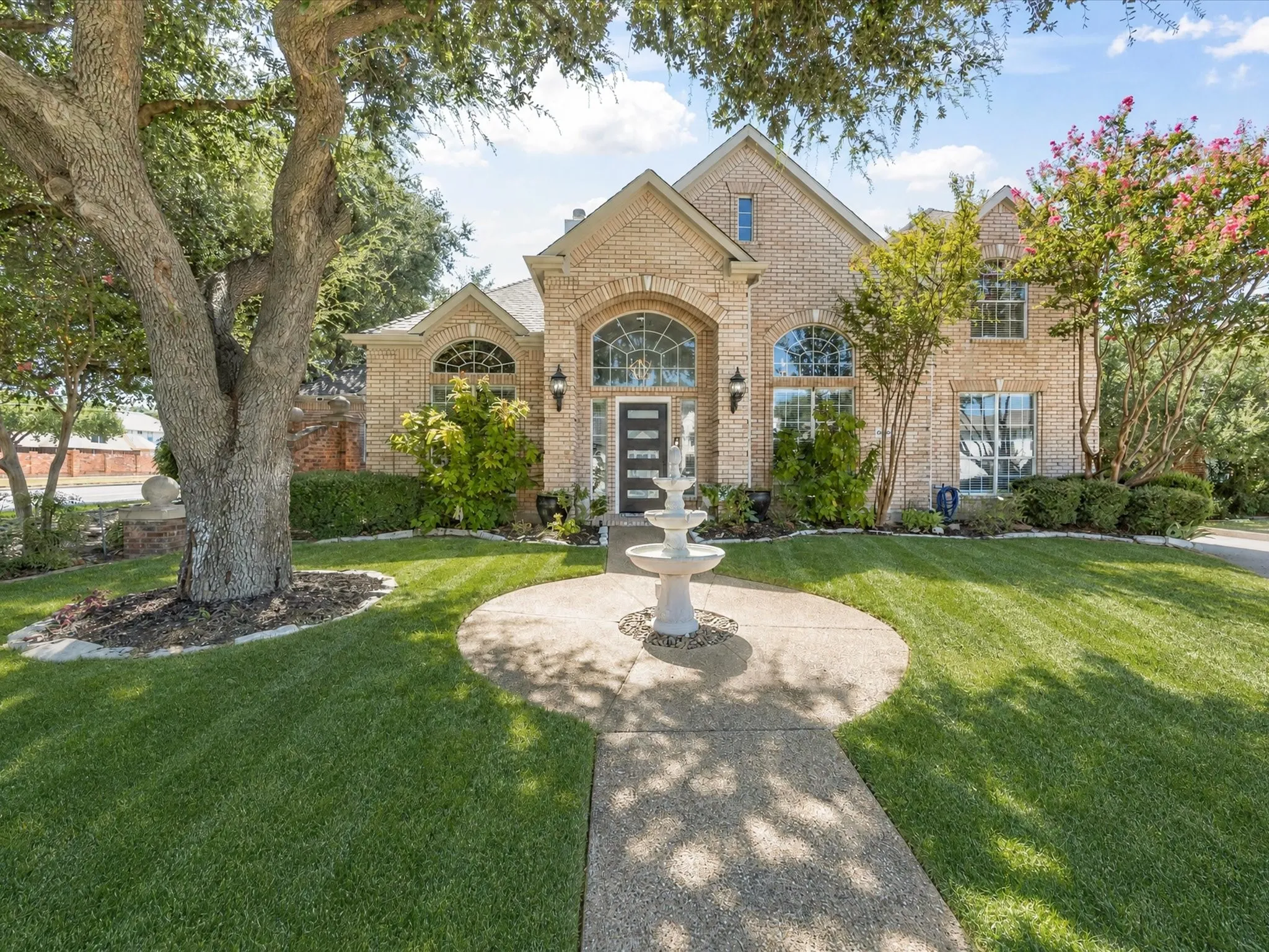 Brick exterior with arched windows, a tiered fountain, mature trees, and a concrete pathway