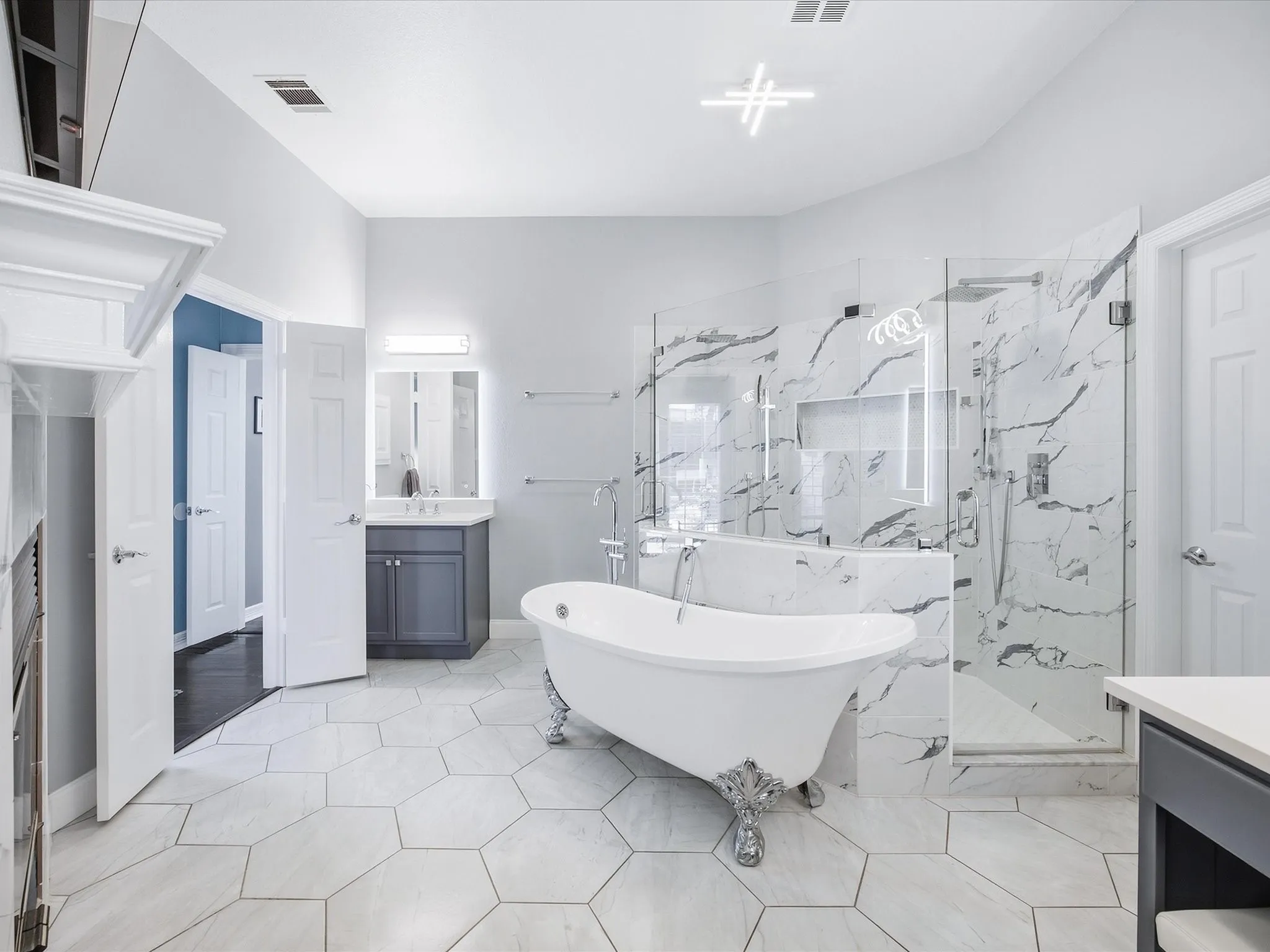 Freestanding soaking tub with ornate feet, glass-enclosed shower with marble-look tile surround, hexagonal floor tiling, double vanity, and contemporary ceiling light fixture