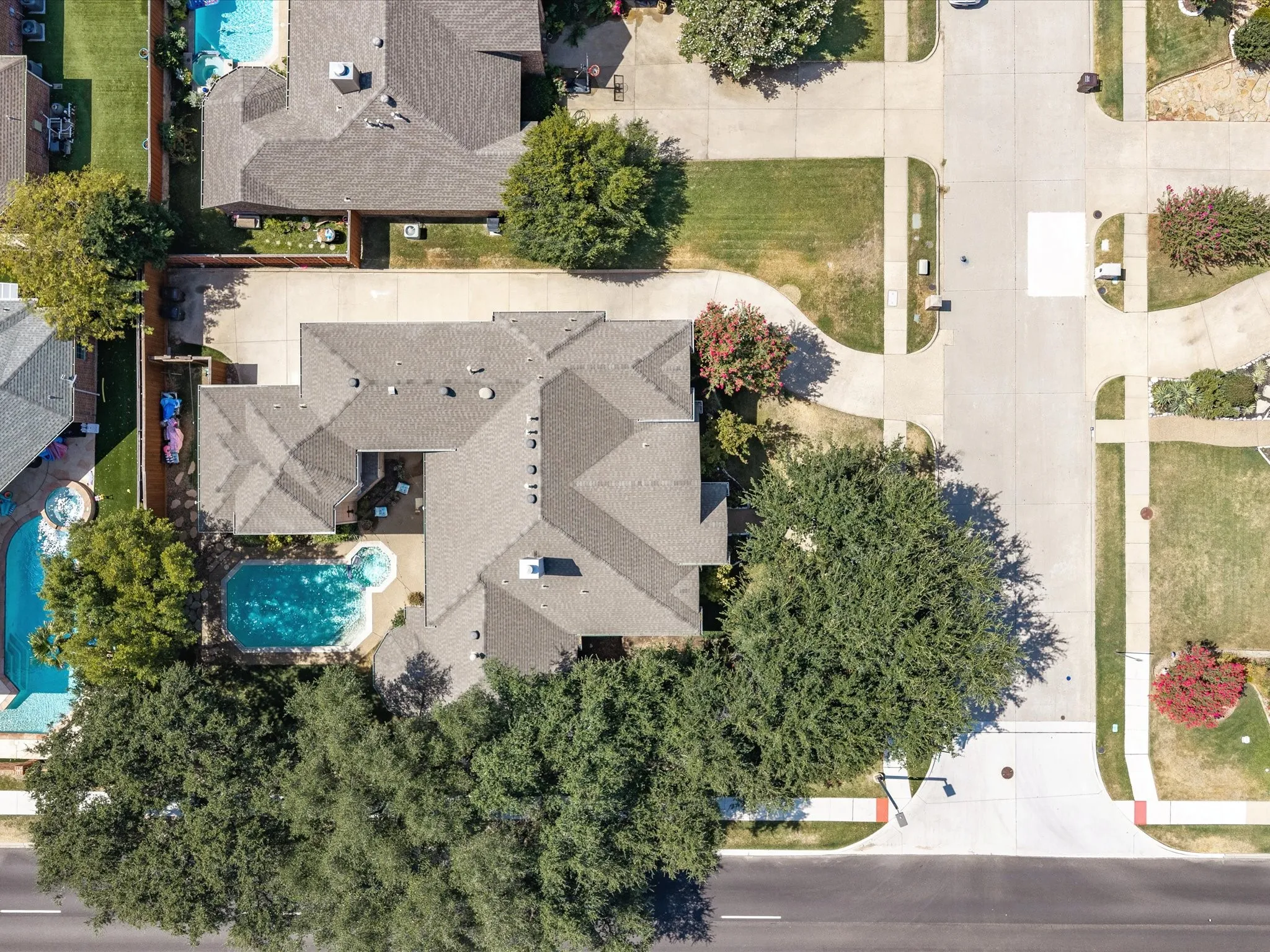 Asphalt shingle roof with multiple gables, extensive paved driveway, established trees, and a sculpted swimming pool with an attached spa