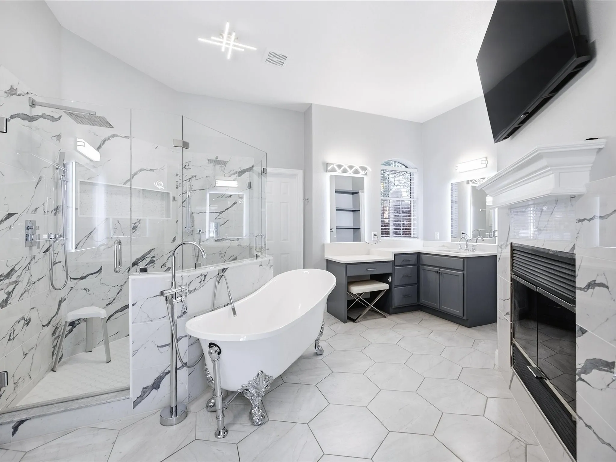 Spacious bathroom featuring a freestanding clawfoot tub, a glass-enclosed shower with a bench, hexagon floor tiling, a dual-sink vanity with gray cabinetry, and a fireplace with a mantel
