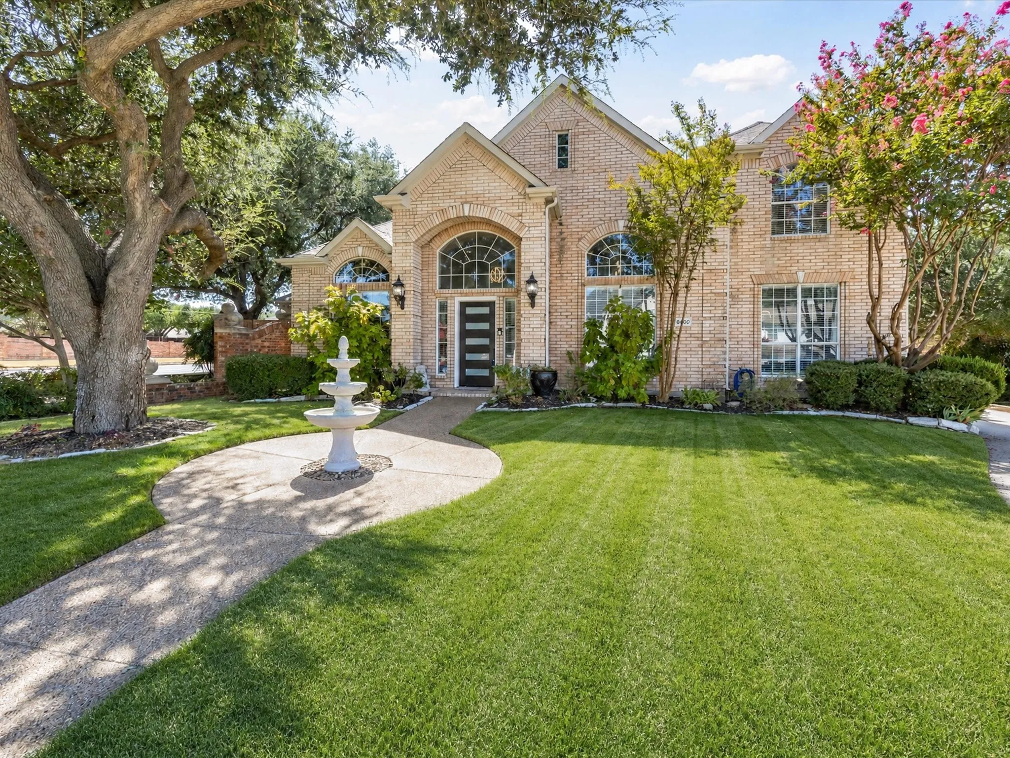 Brick facade with arched windows, mature trees, tiered fountain, and a landscaped front yard