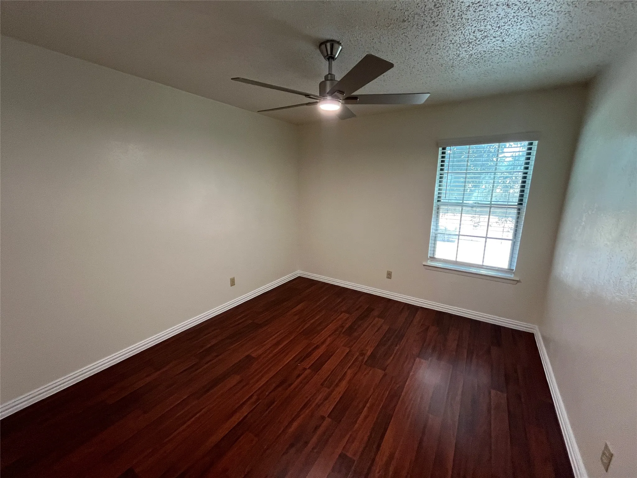 Rich wood-finish flooring, single window with blinds, ceiling fan with integrated lighting, and neutral wall tones