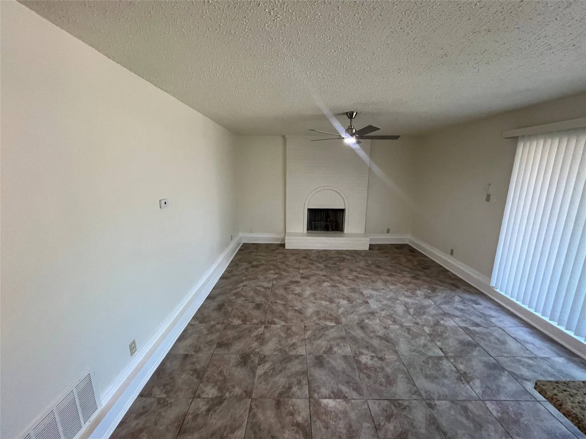 Spacious room featuring tile flooring, white brick fireplace, modern ceiling fan, and vertical window blinds