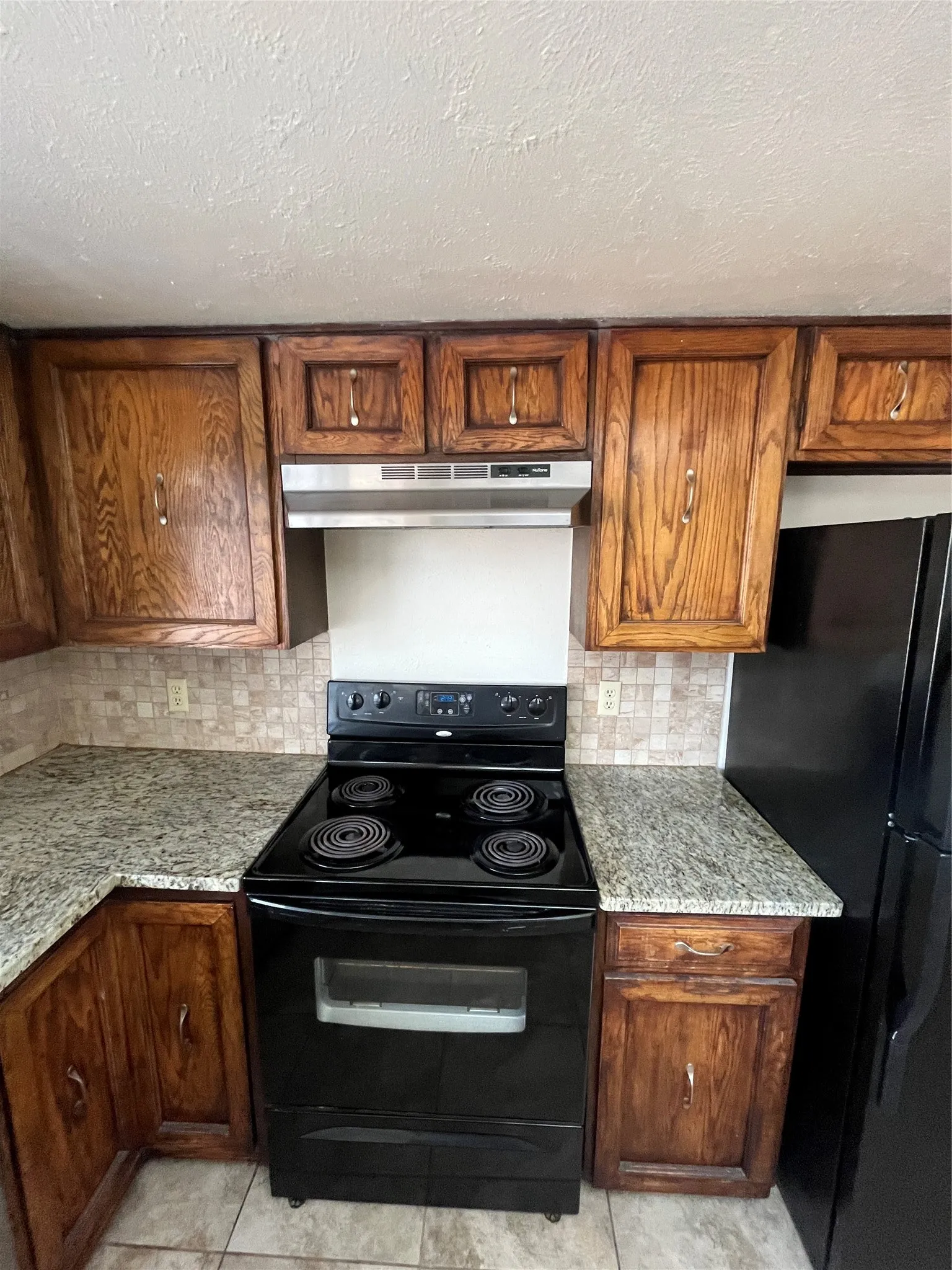 Wood cabinetry, tile backsplash, black electric range, and granite-style countertops