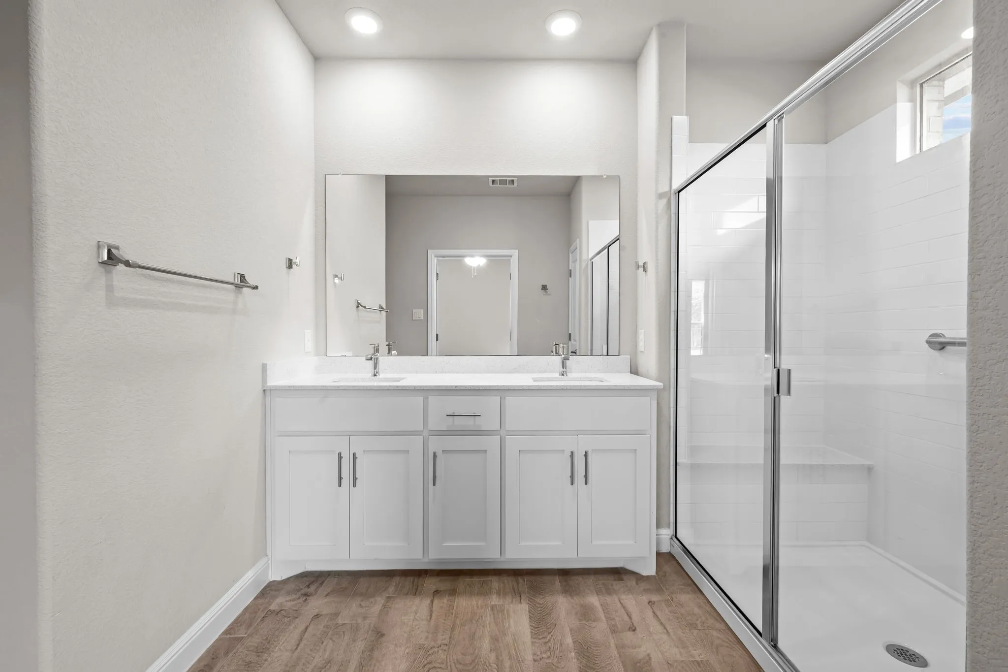 Dual vanity with white cabinetry, wood-finish flooring, a frameless glass shower enclosure, and a built-in shower bench