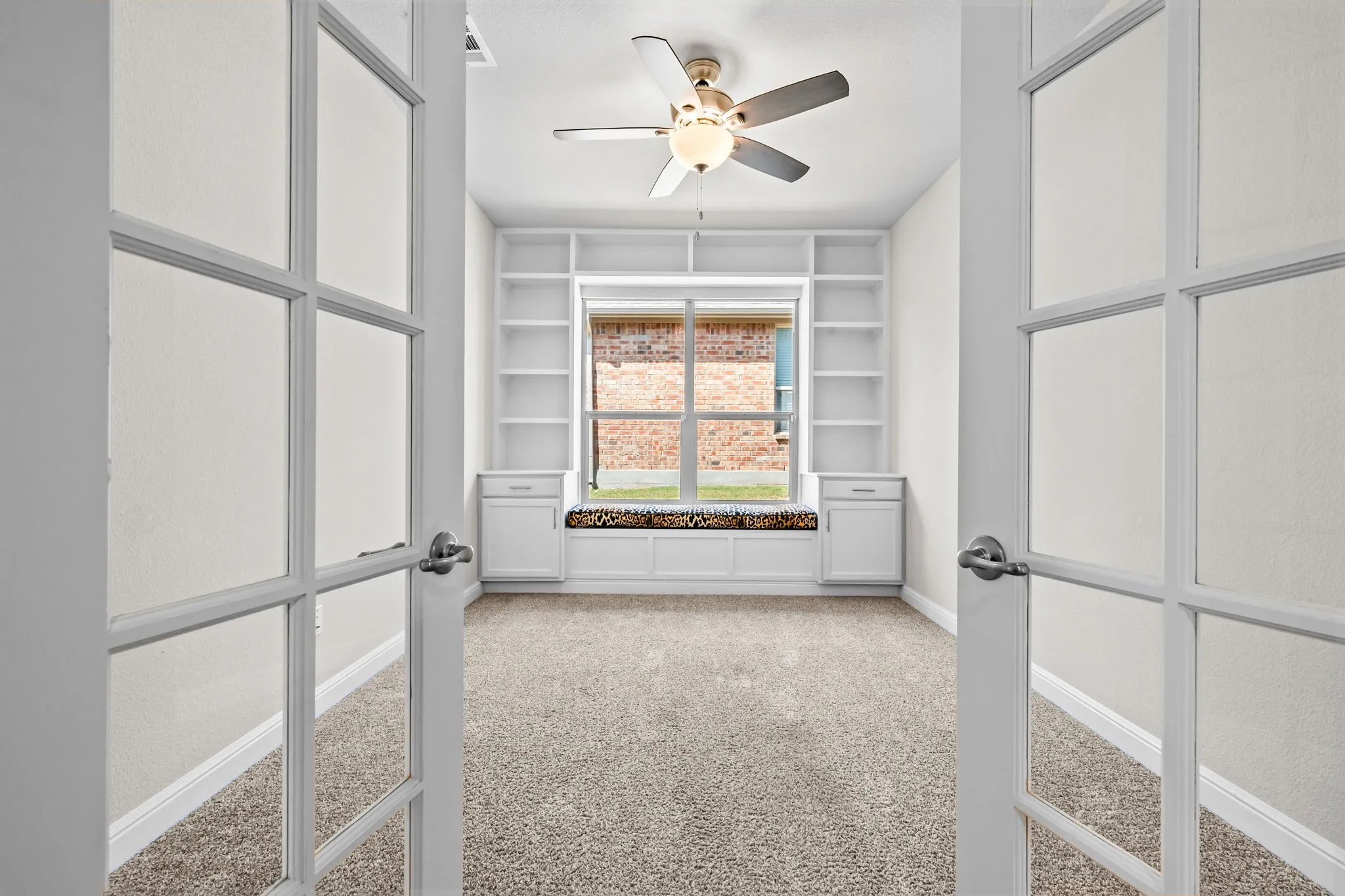 French doors opening to a versatile room with carpet flooring, built-in shelving, a window seat with storage, and a ceiling fan with integrated lighting
