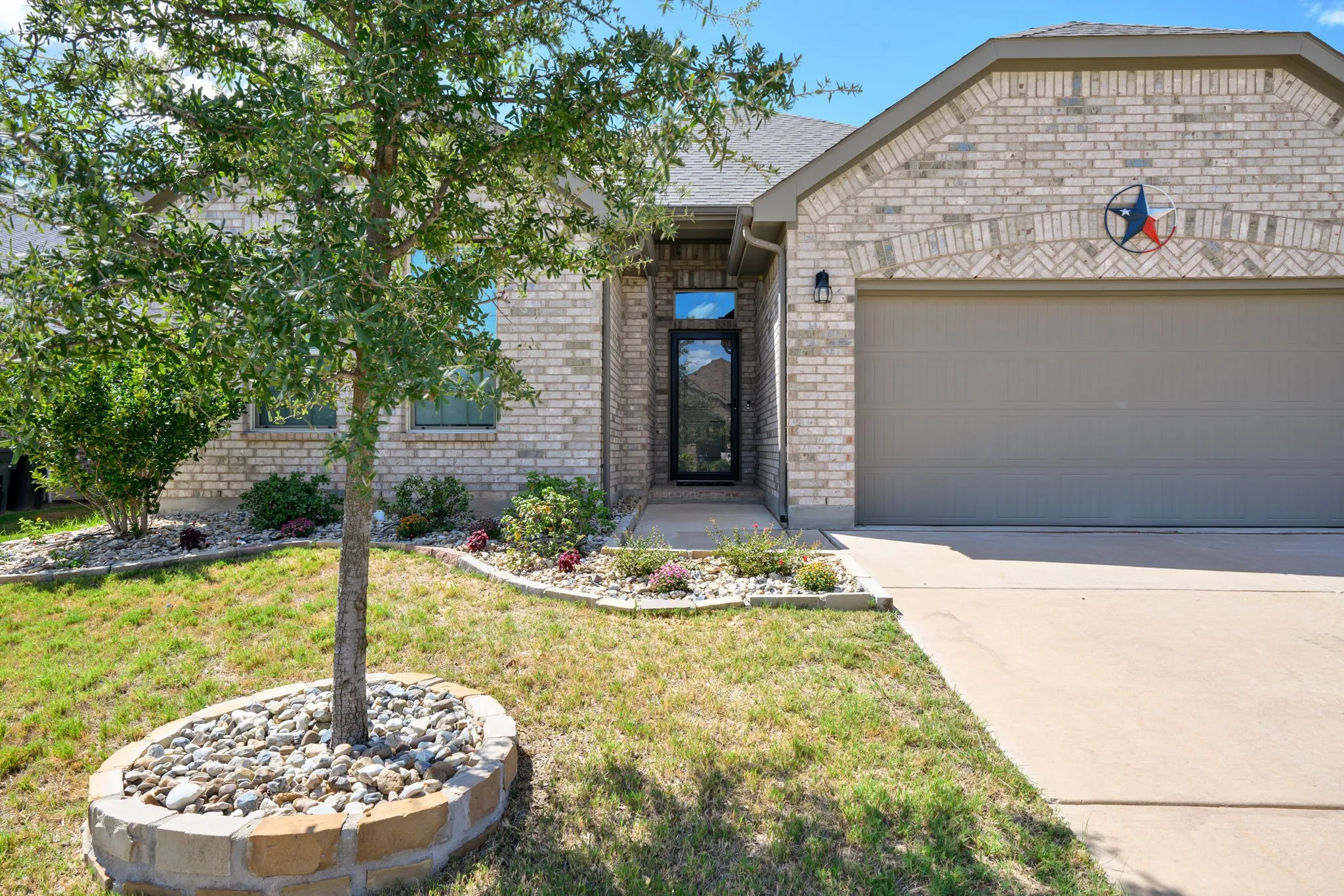 Light-colored brick exterior, attached garage, concrete driveway, front entry door with transom window, and mature landscaping