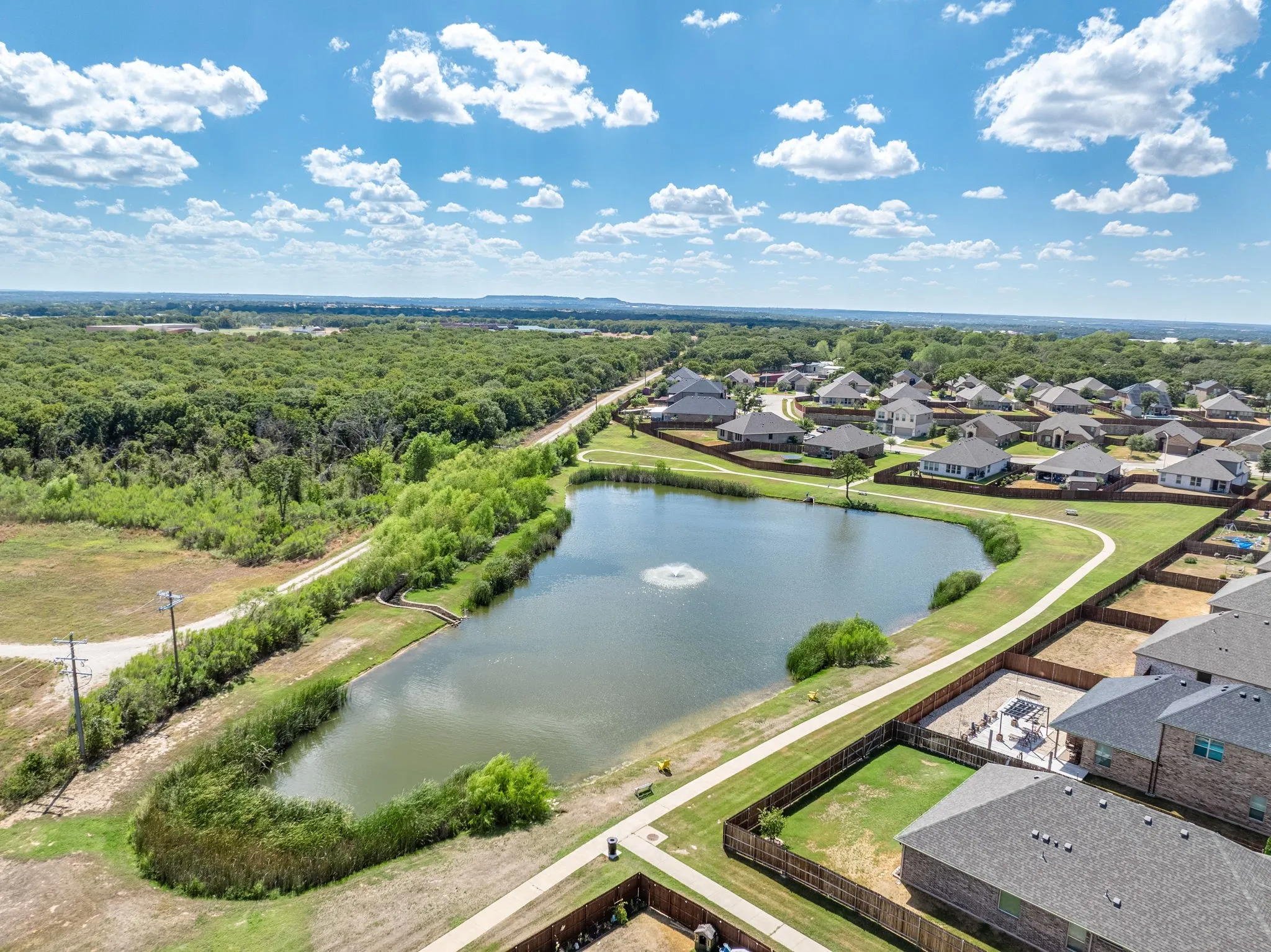 Neighborhood setting featuring a large pond with a fountain, a paved walking path, and a surrounding expanse of mature trees