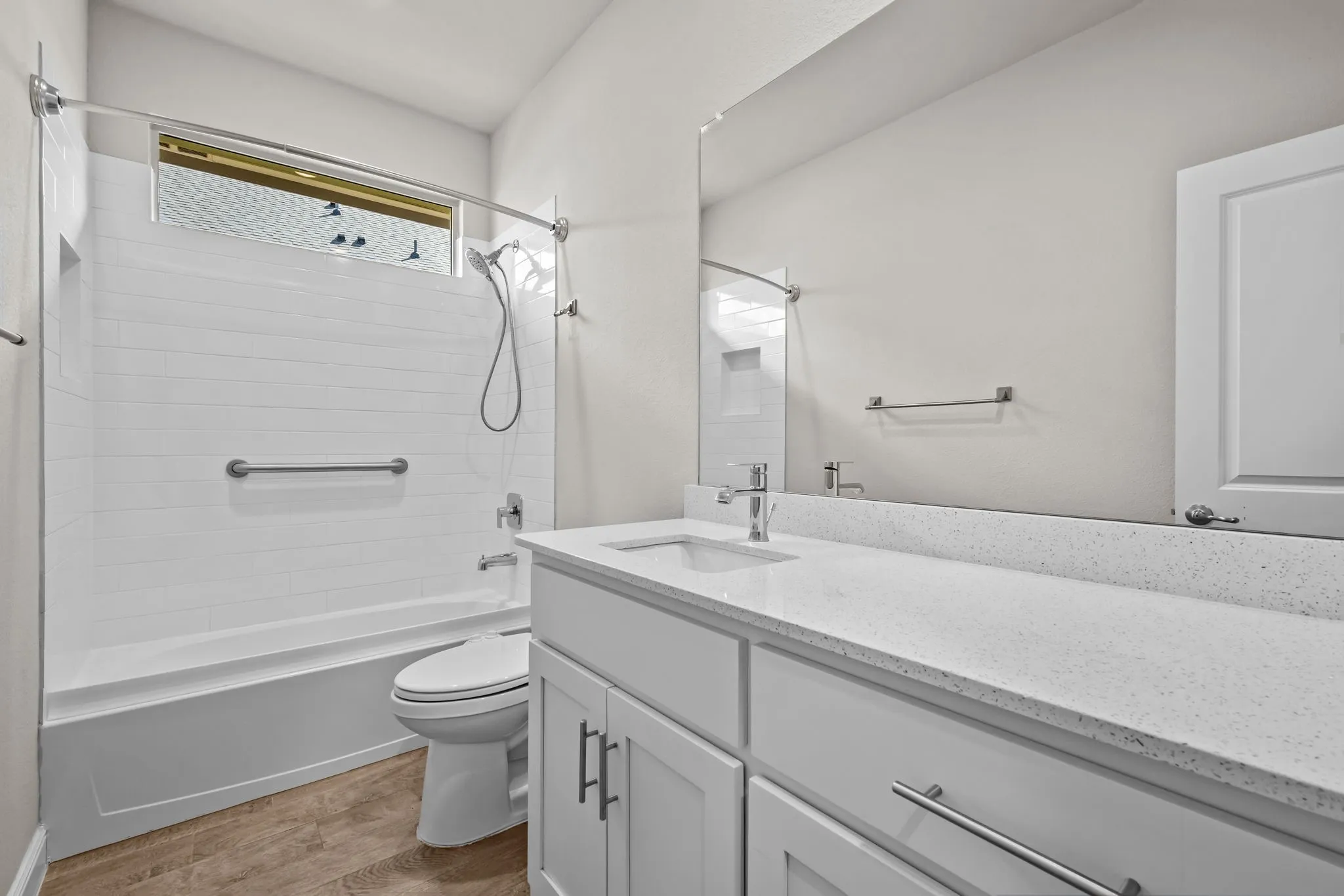 White vanity with a light-toned countertop, wood-finish flooring, and a white tiled shower/tub combination with a window