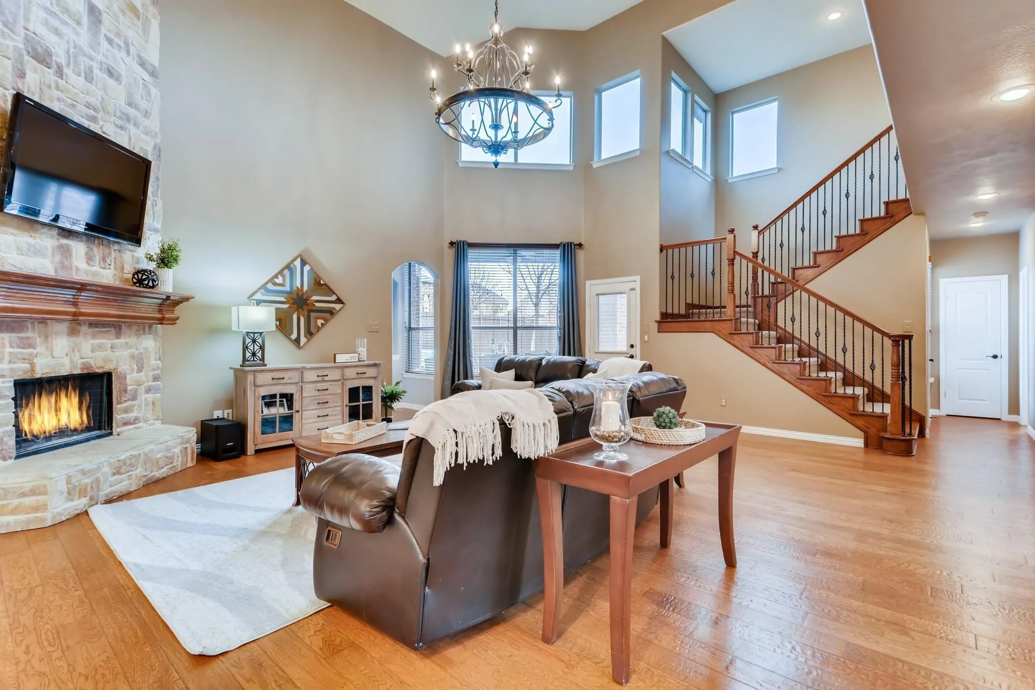 Two-story living area with high ceilings, stone fireplace, a grand staircase with wood treads and iron balusters, and wood-finish flooring throughout
