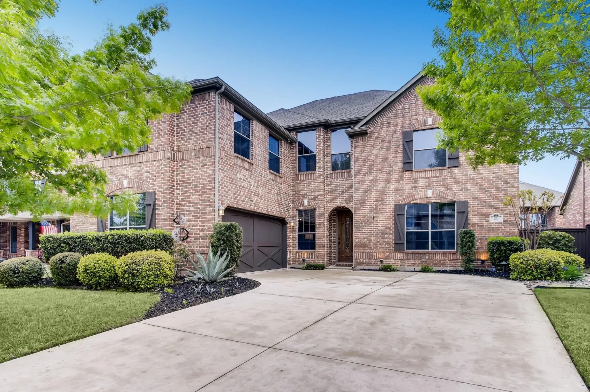 Brick exterior with dark shutters, arched entry, two-car garage, concrete driveway, and manicured landscaping