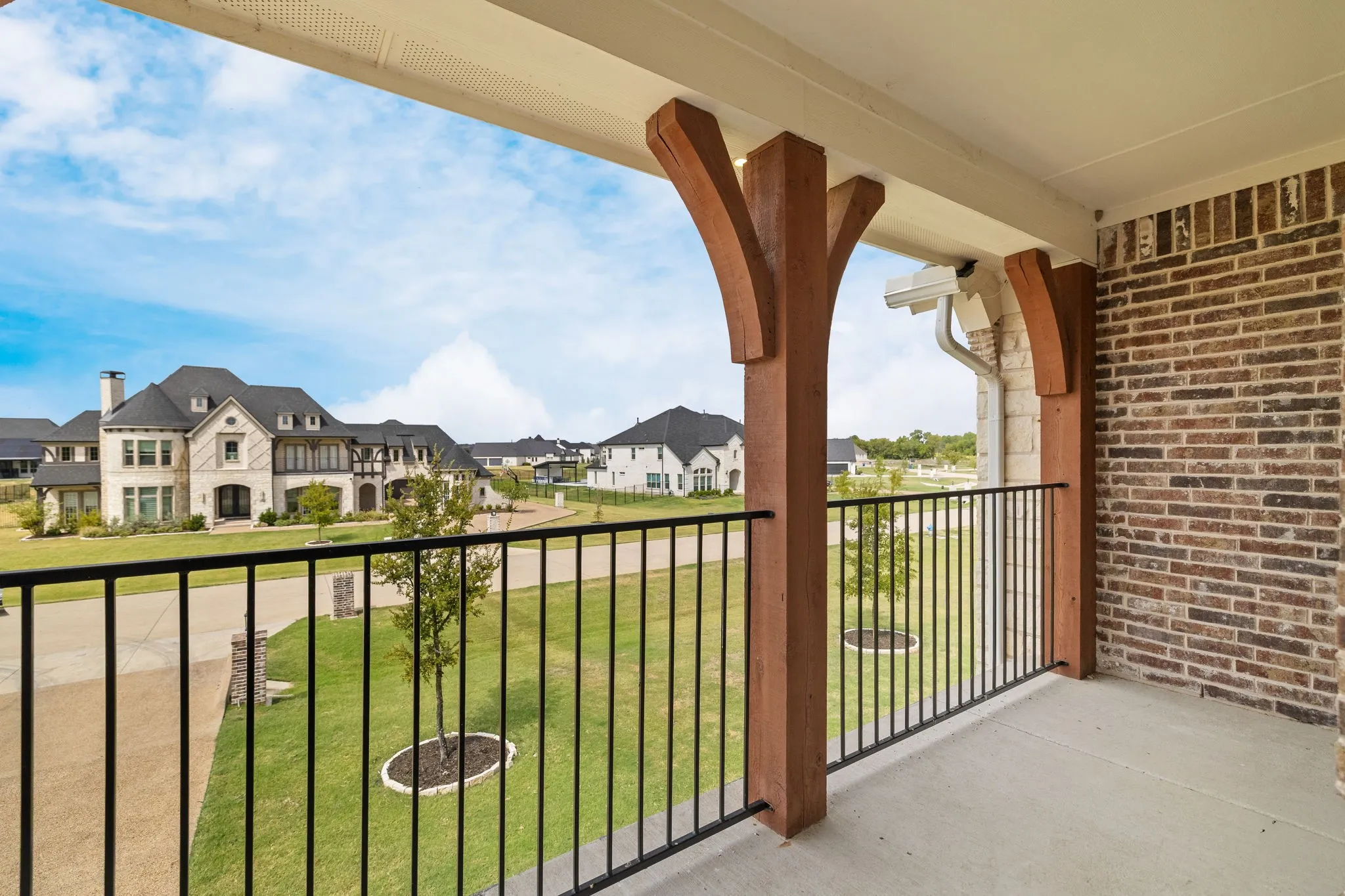Covered balcony featuring wood-finish support beams, wrought iron railing, concrete flooring, and brick siding