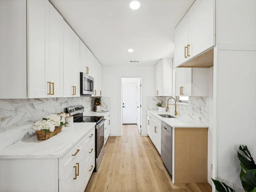 Galley kitchen featuring abundant white cabinetry with gold-finish hardware, stainless steel appliances, light-toned countertops, and a full-height backsplash