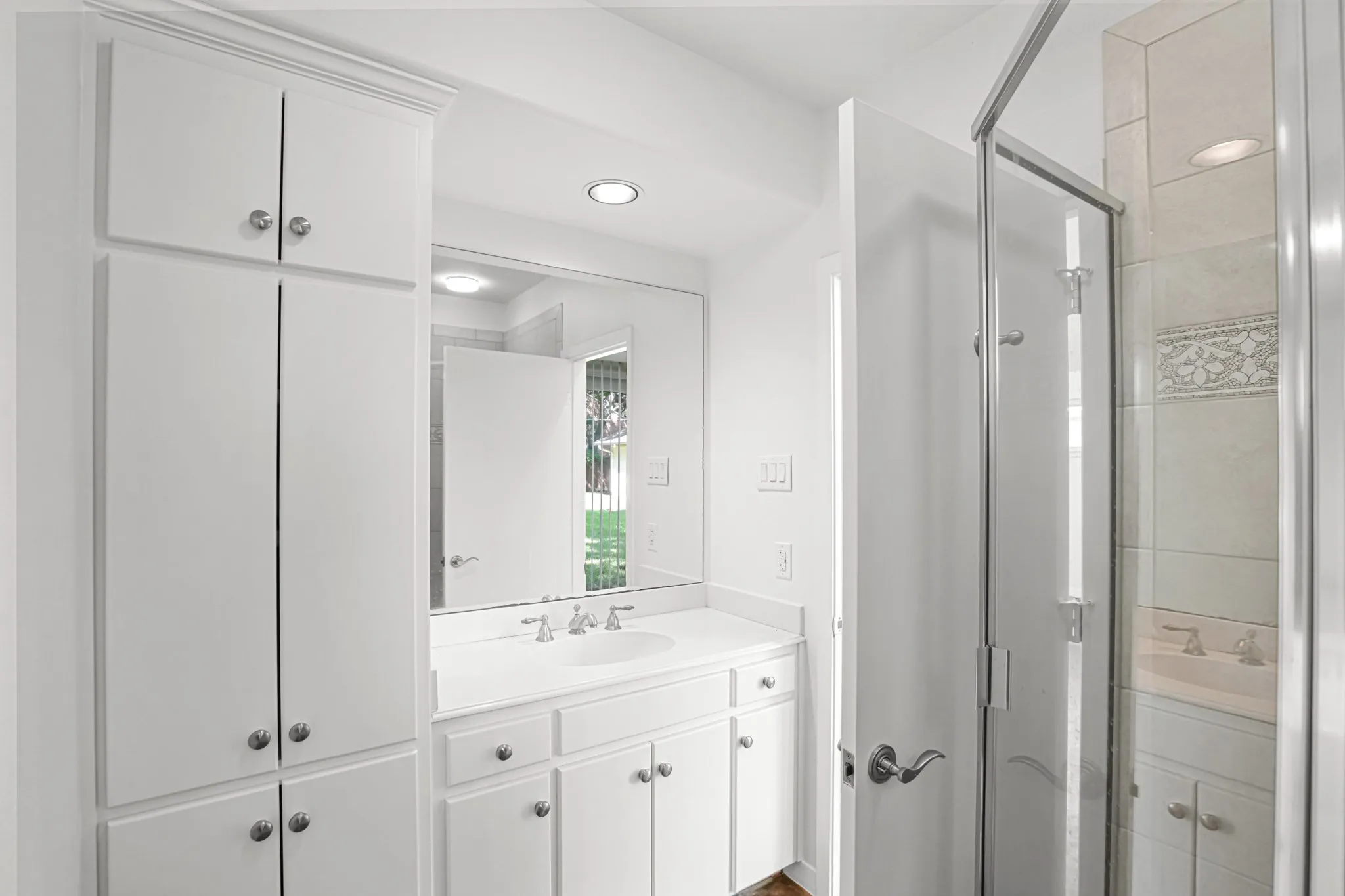 White cabinetry with chrome hardware, single basin vanity, large vanity mirror, glass-enclosed shower, and tiled shower surround with decorative accents.