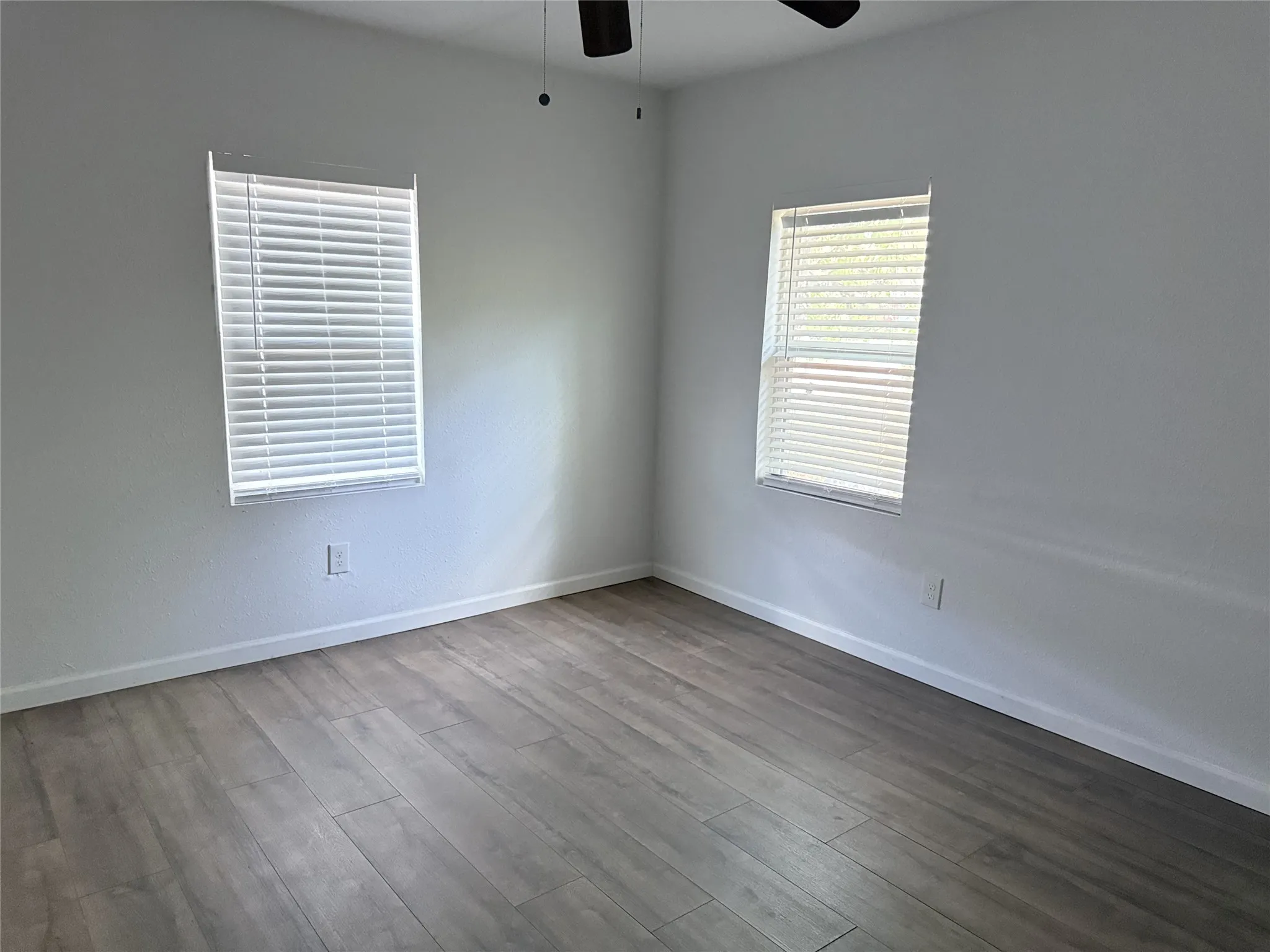 Wood-finish flooring, white walls, two windows with blinds, and a ceiling fan with integrated lighting